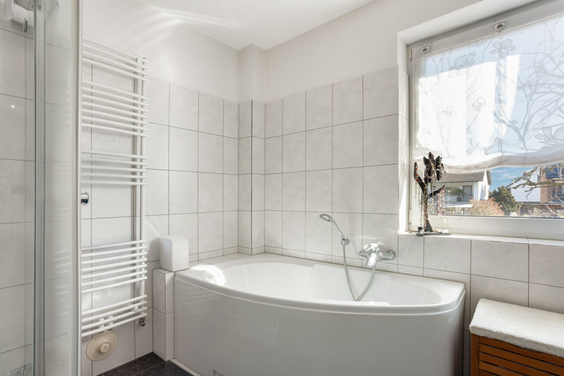 Helles Badezimmer mit Eckbadewanne, Handtuchheizkörper und Fenster mit Vorhang.