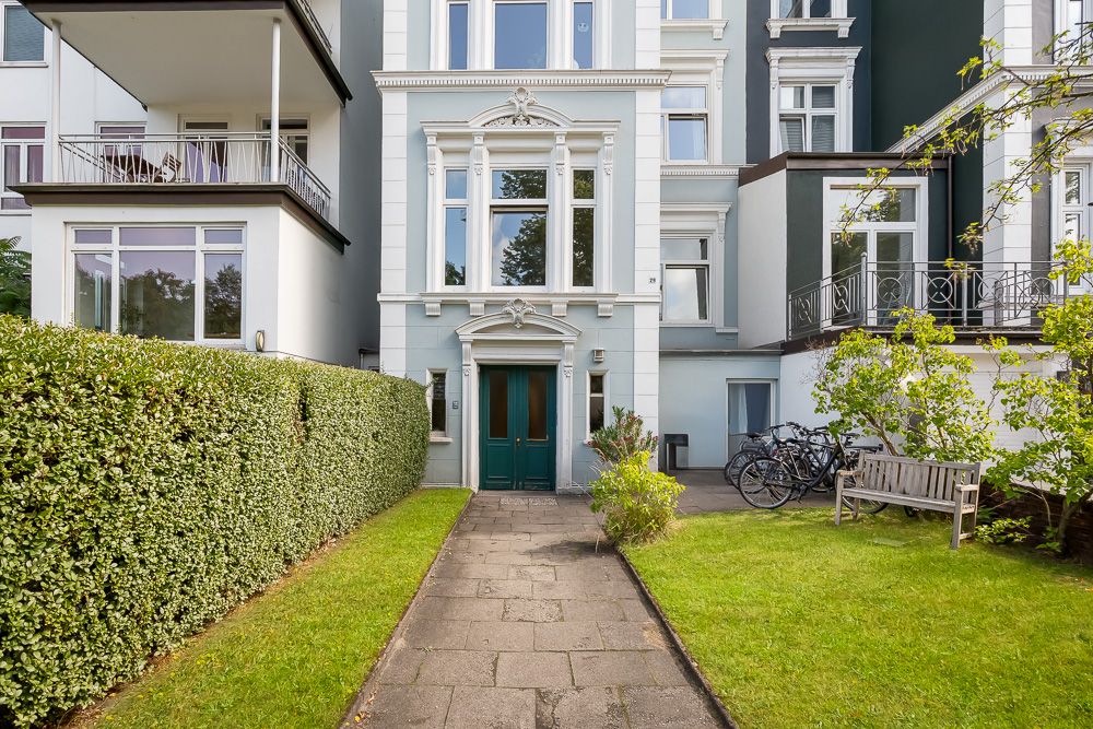 Eingang eines Wohnhauses mit grünem Vorgarten, Bank, Hecke und Fahrrädern.