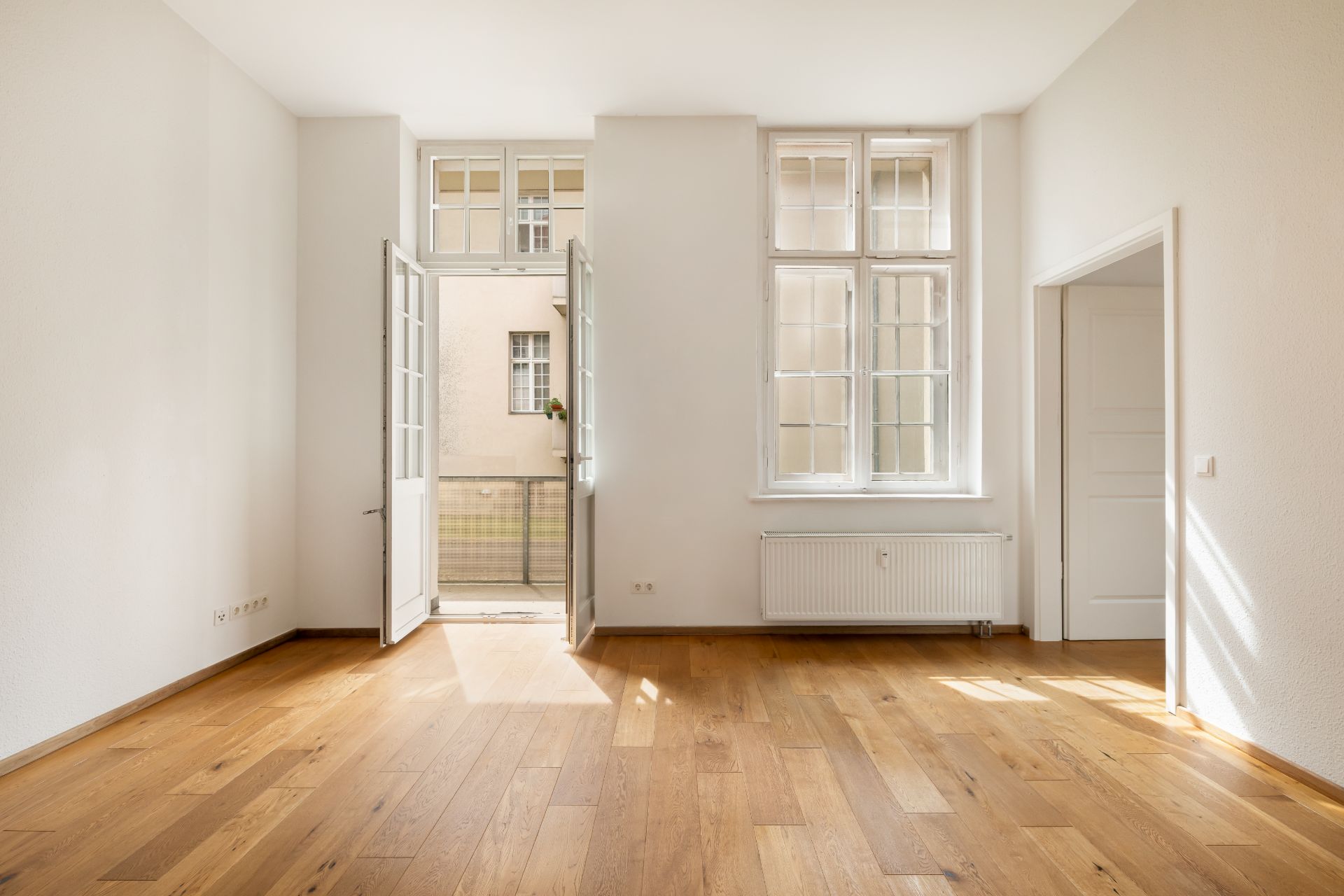 Helles Zimmer mit Holzboden, großen Fenstern, Tür zum Balkon und offenem Durchgang.