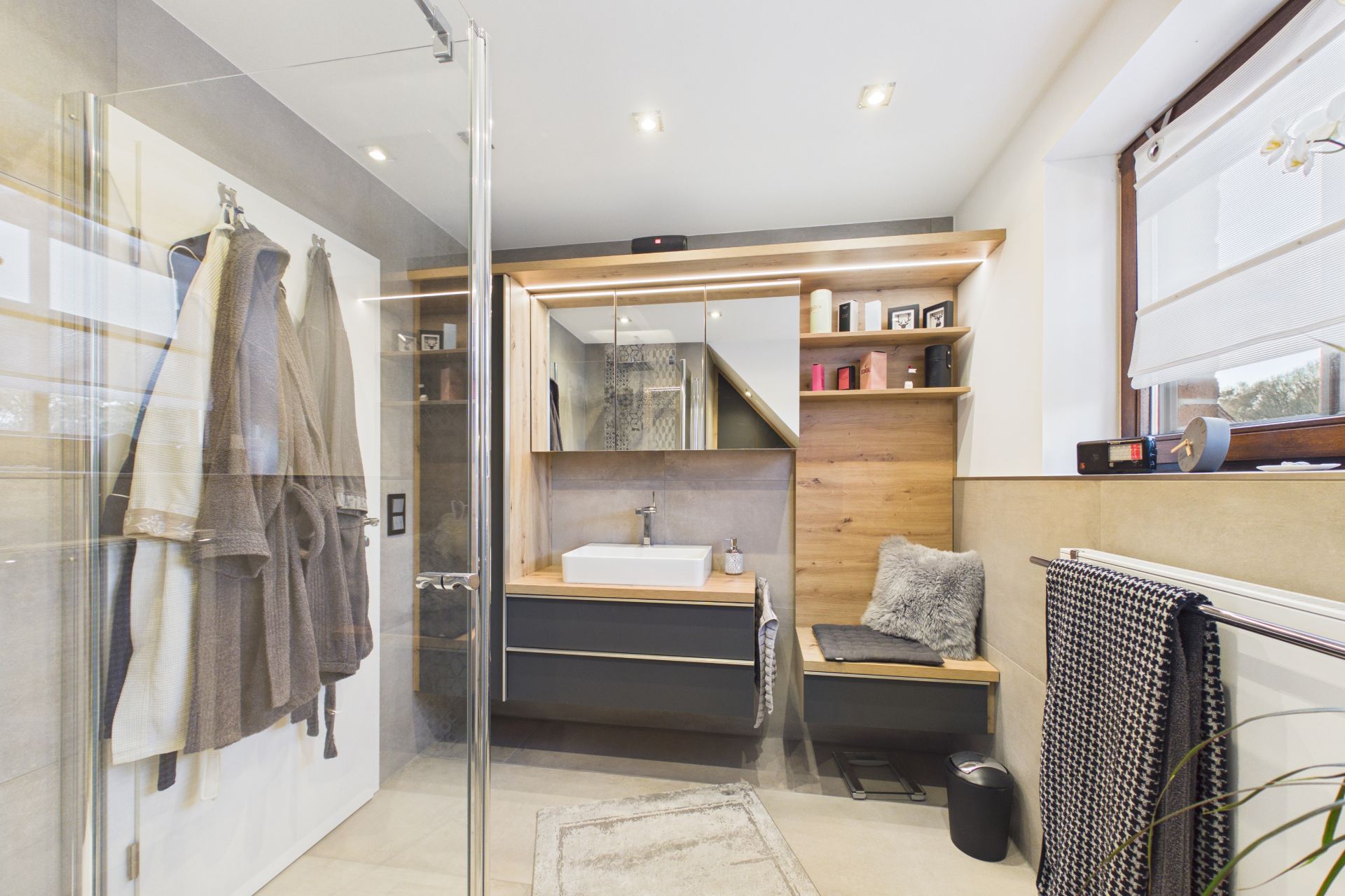 Modernes Badezimmer mit Waschbecken, Spiegel, Dusche und Handtüchern, helle Einrichtung und Fenster.
