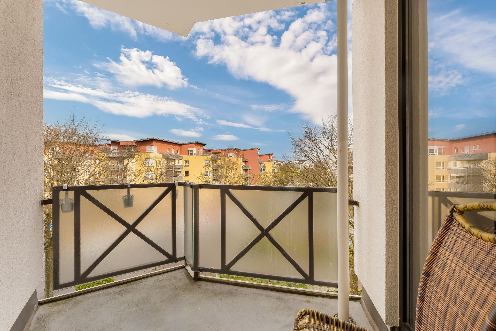 Balkon mit Glasgeländer und Blick auf Wohngebäude bei blauem Himmel.