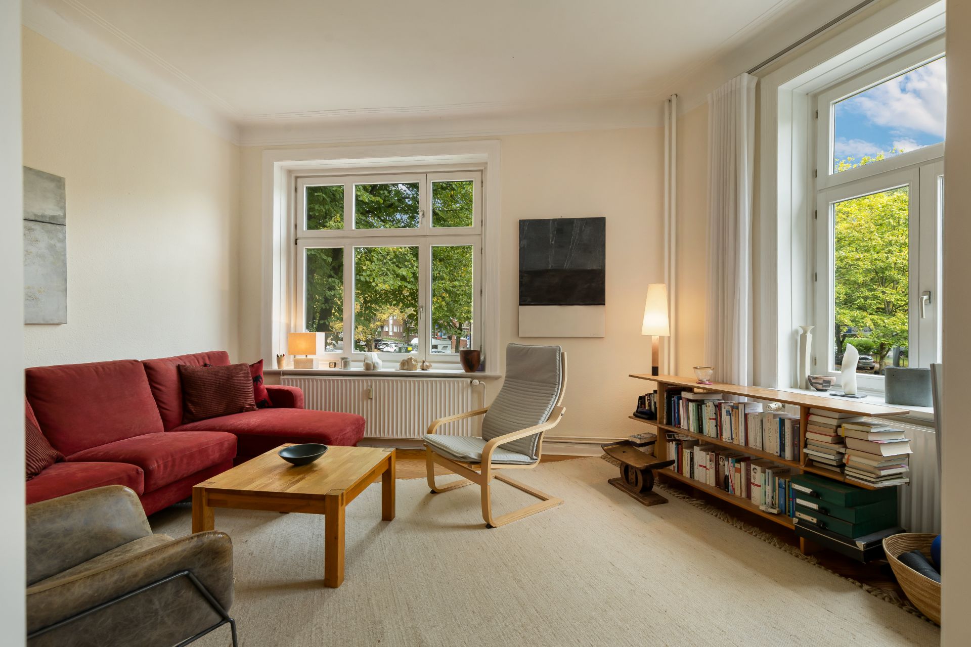 Helles Wohnzimmer mit rotem Sofa, Sessel, Holztisch und Bücherregal am Fenster.