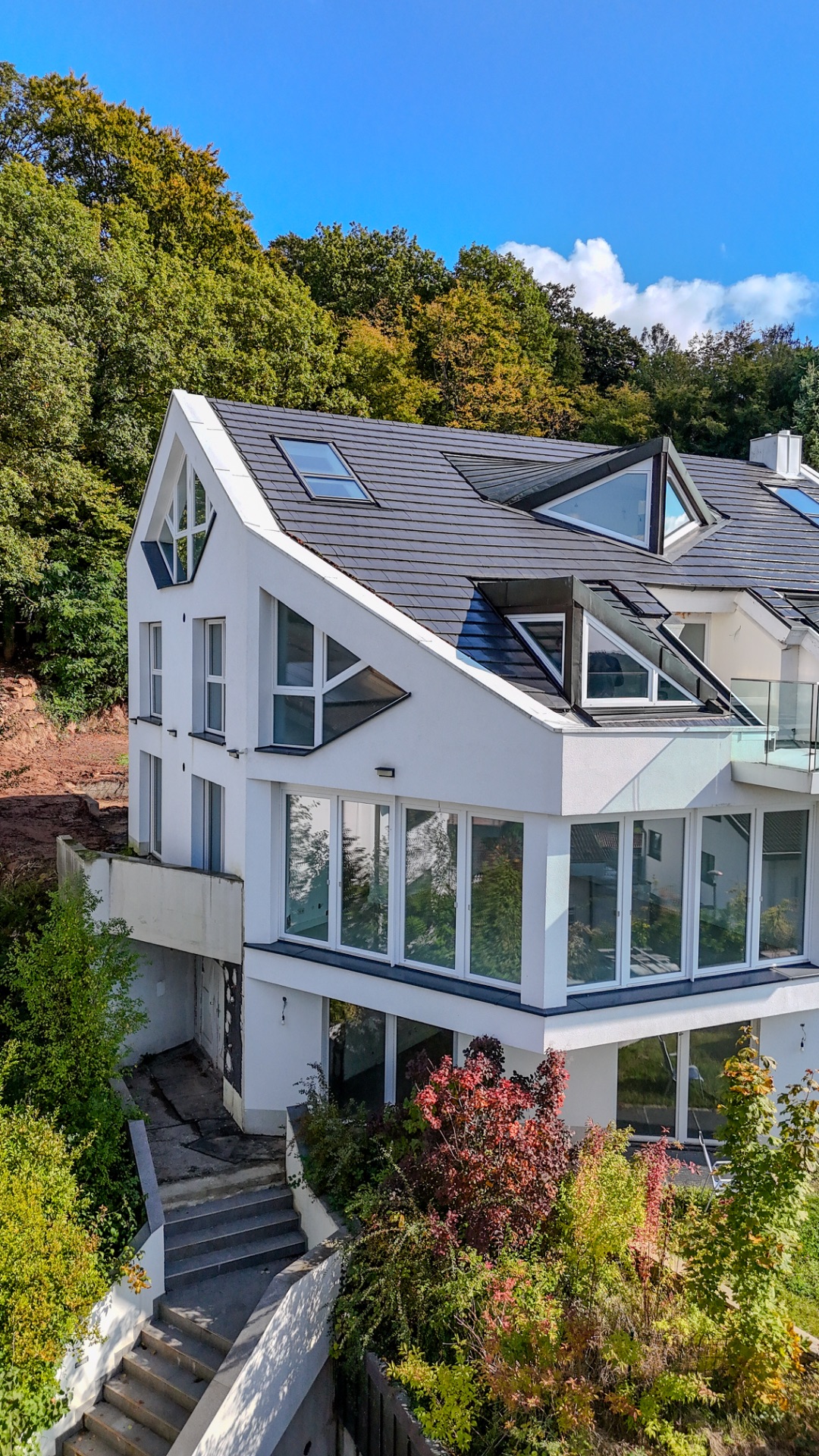 Modernes weißes Wohnhaus mit großen Fenstern und dunklem Schrägdach, umgeben von Bäumen.