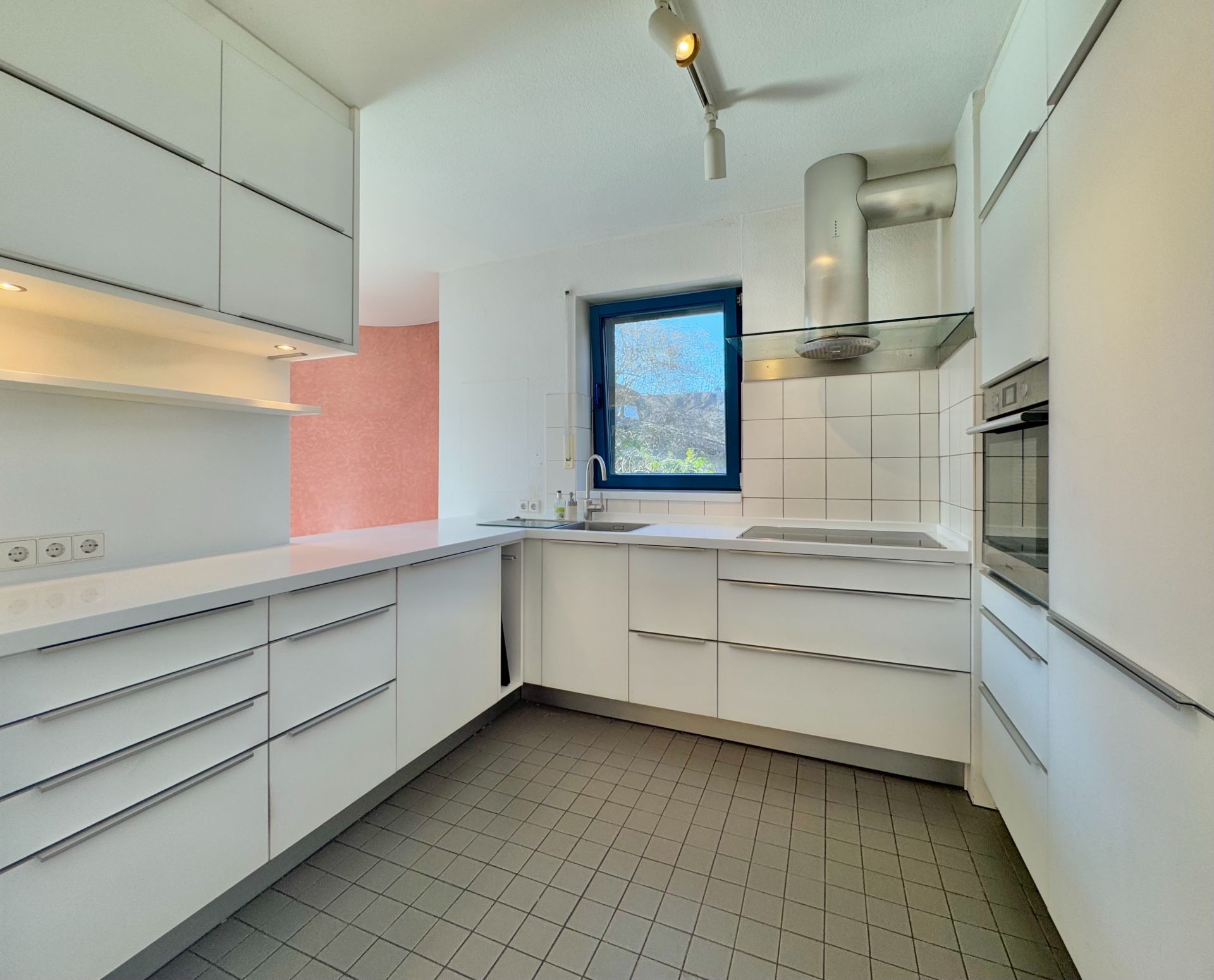 Moderne weiße Einbauküche mit Fenster, Kochfeld, Backofen und vielen Schränken.