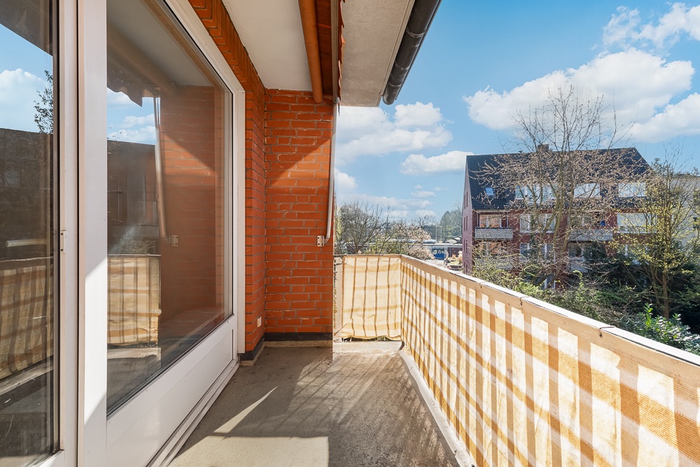 Balkon mit Backsteinwand und Geländer, daneben große Glastür, Blick auf Nachbarhäuser und Bäume.