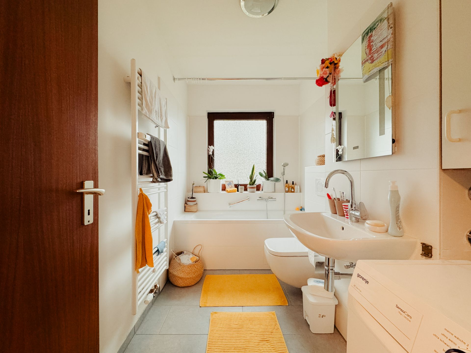 Helles Badezimmer mit Badewanne, Waschbecken, WC, Waschmaschine und gelben Badvorlegern.