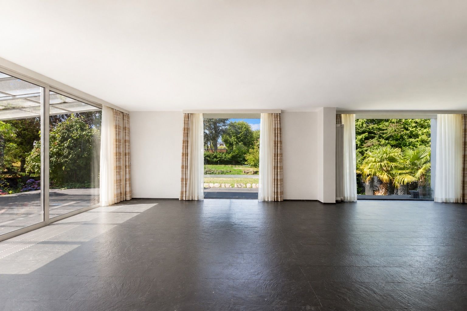Heller leerer Raum mit großen Fenstern und Blick auf einen Garten mit Bäumen.