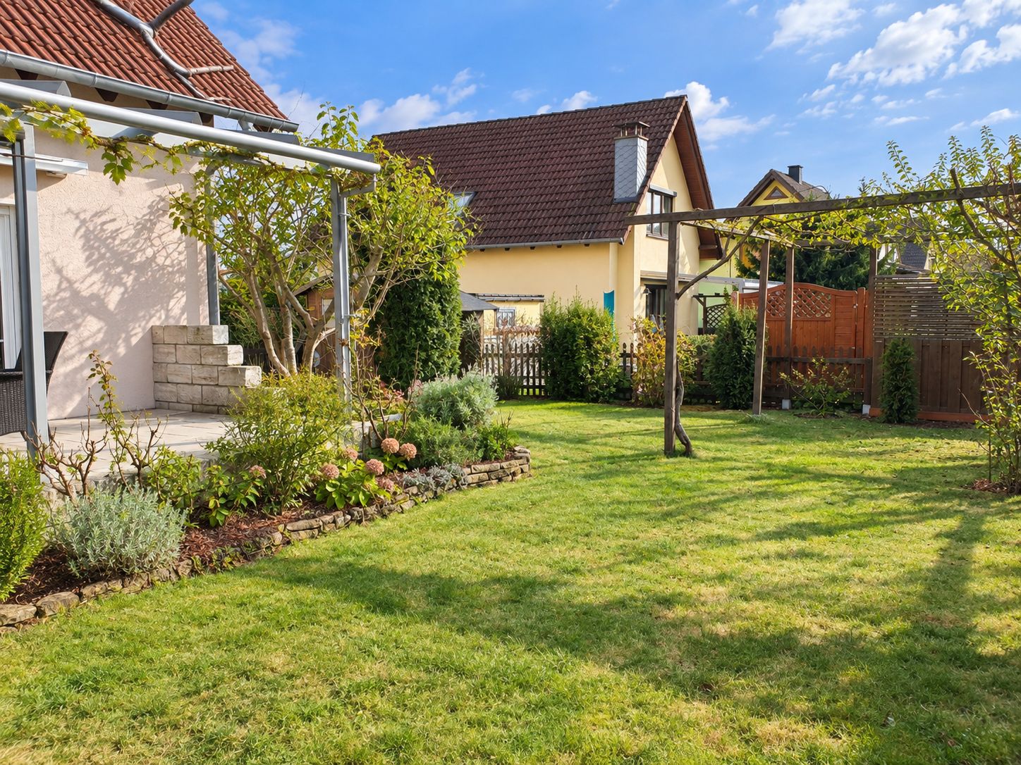 Grüner Garten mit Beeten, Rasen und Pergola neben einem Wohnhaus.