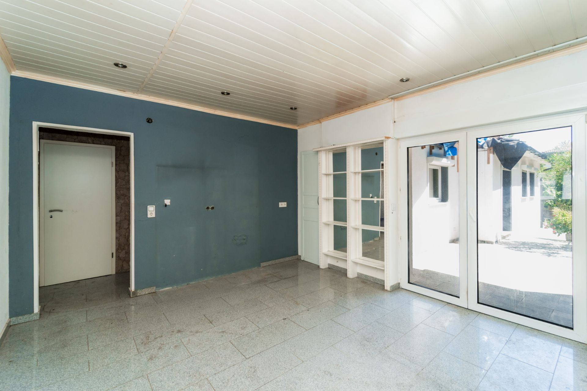 Heller Raum mit blauer Wand, Glastür, weißen Regalen und Blick in den Garten.