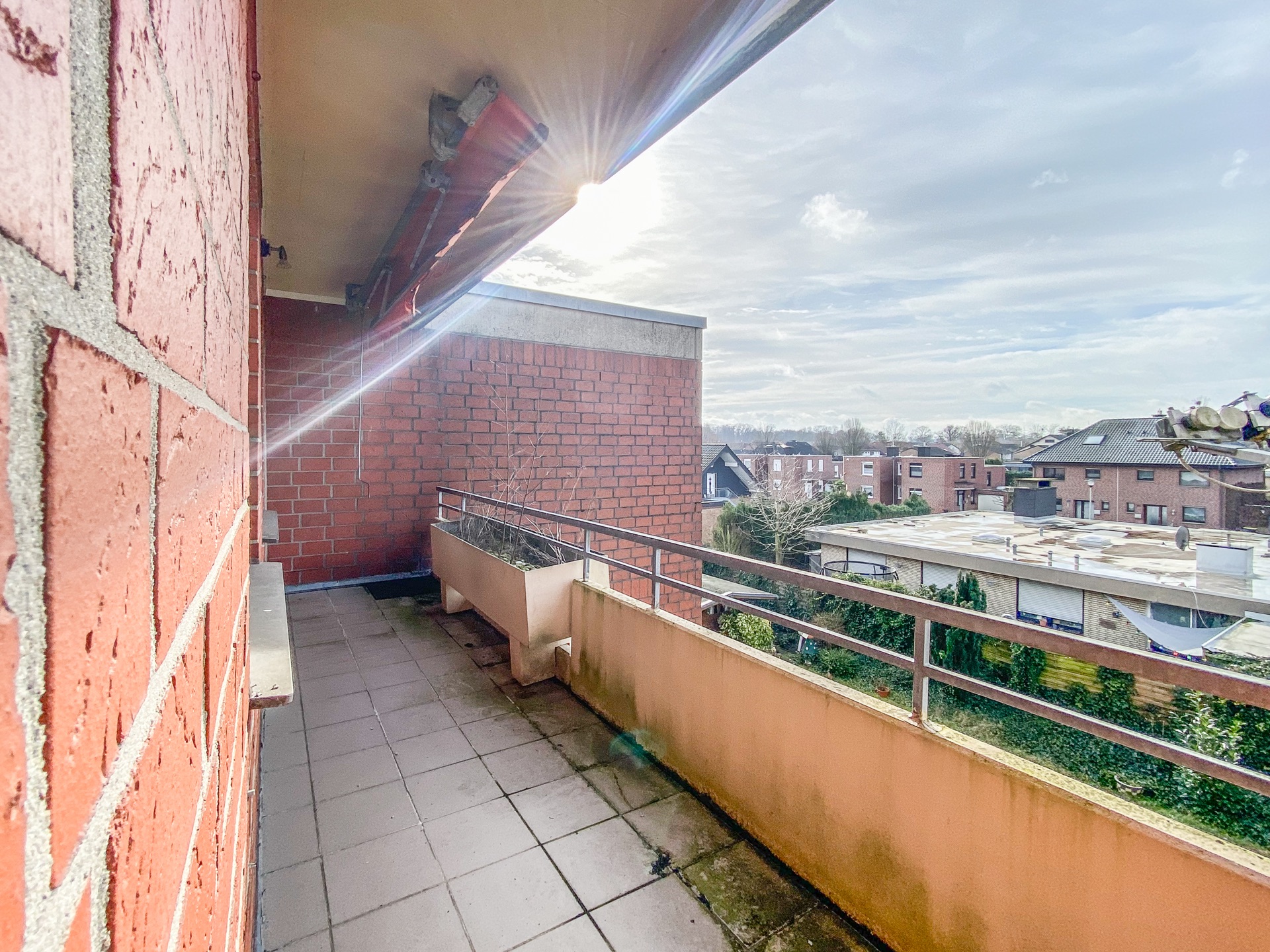 Balkon mit Backsteinwand, Geländer und Pflanzenkasten, Ausblick auf Wohnhäuser.