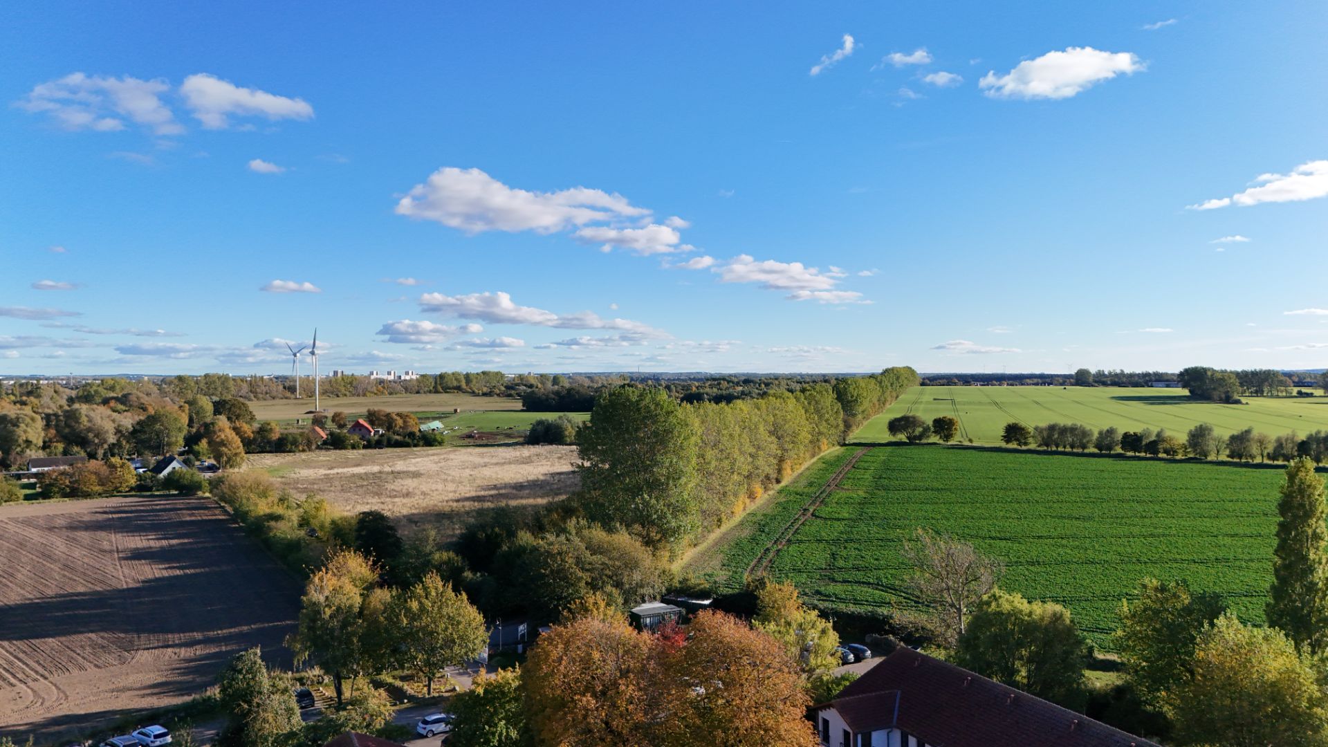 Blick über Felder, Bäume und Häuser unter blauem Himmel mit einigen Wolken.