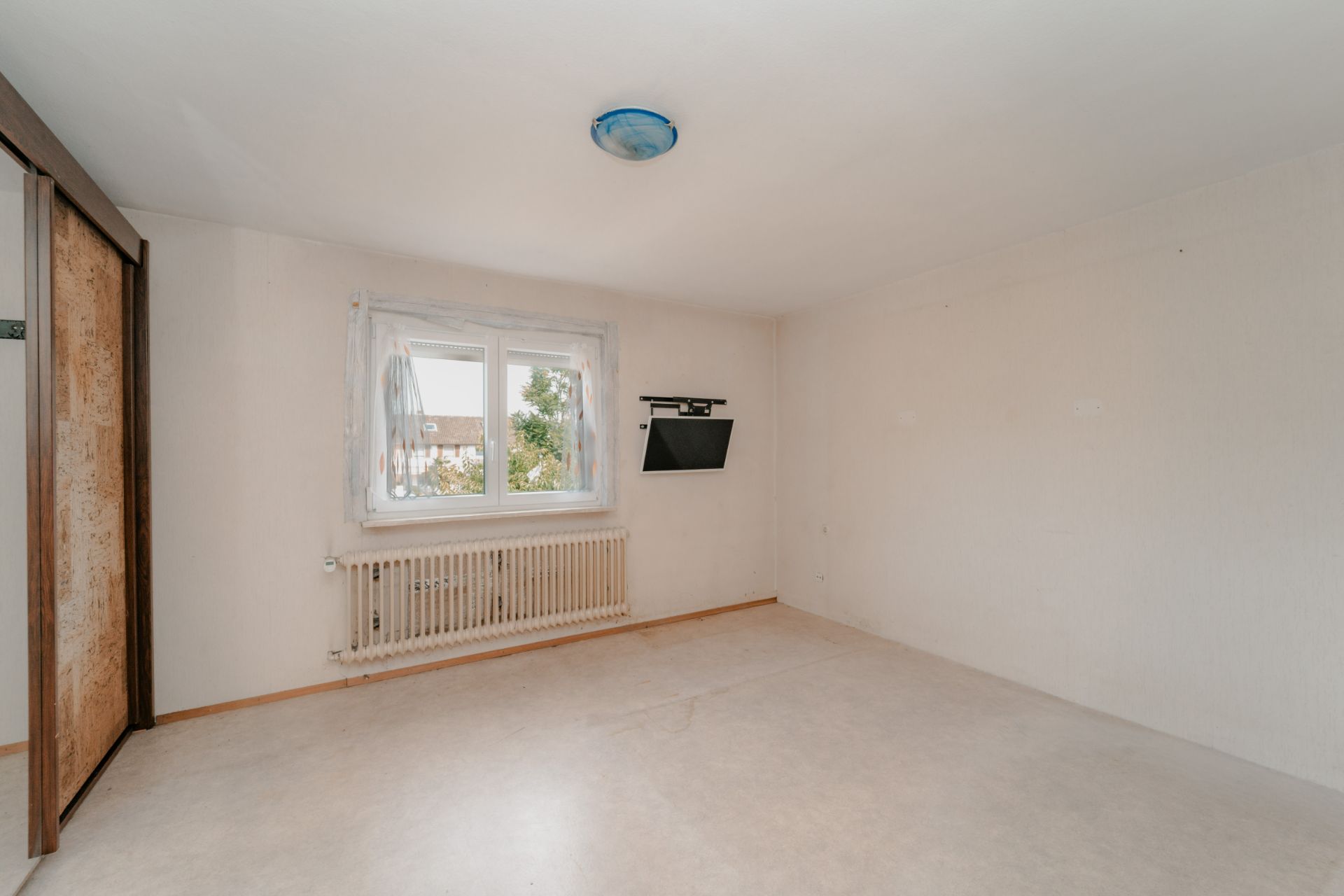 Leeres Zimmer mit Fenster, Heizkörper, Wandregal und großem Schrank mit Schiebetüren.