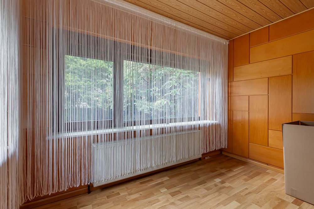 Leerer Raum mit Holzverkleidung, großem Fenster und weißen Fadengardinen.