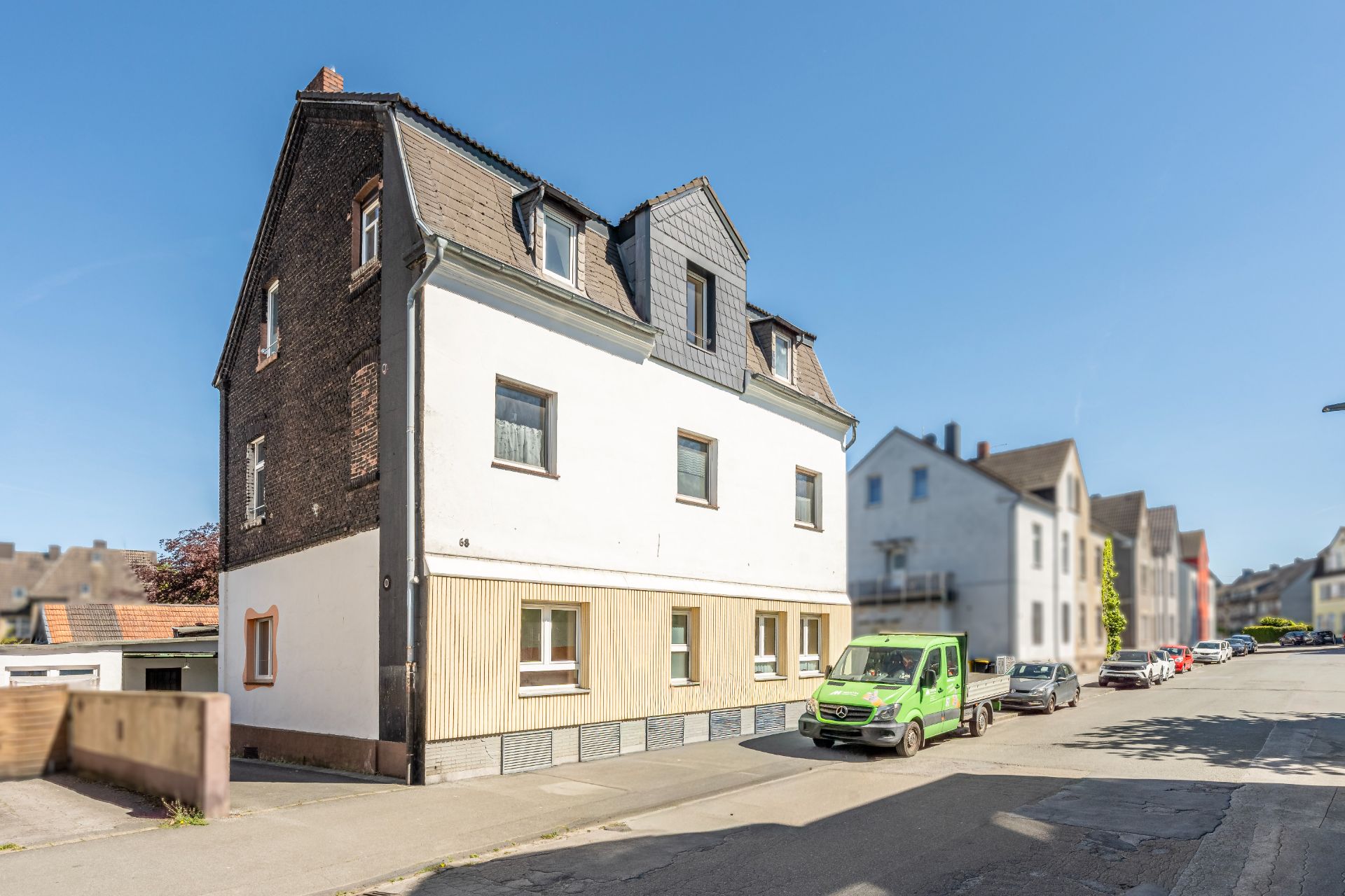 Dreistöckiges Wohnhaus mit dunklem Dach an einer ruhigen Straße, mehrere geparkte Autos sind zu sehen.
