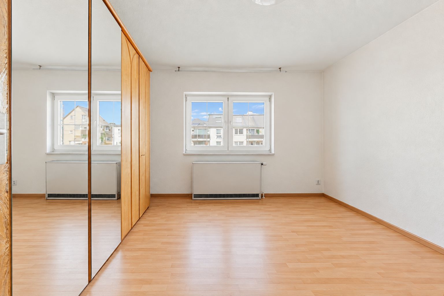 Helles, leeres Zimmer mit Holzfußboden, zwei Fenstern und einem großen Schrank mit Spiegeltüren.