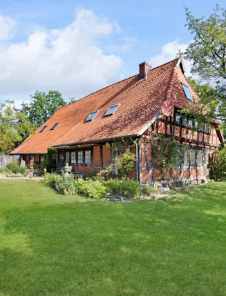 Großes Fachwerkhaus mit rotem Dach und Garten, Bäume und Wiese im Vordergrund.