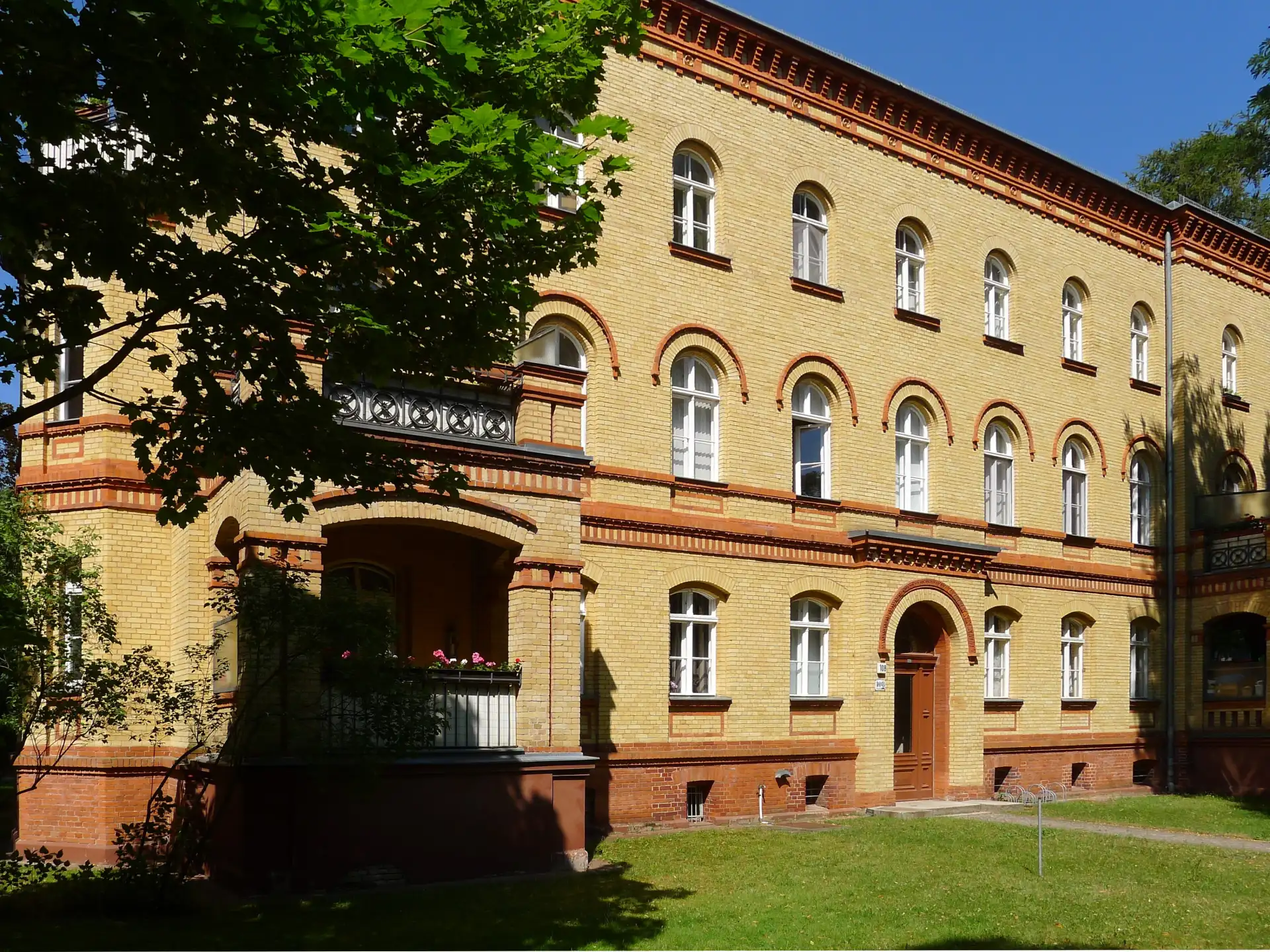 Dreistöckiges Wohnhaus aus gelbem Klinker mit weißen Fenstern und Balkon, davor Rasenfläche.