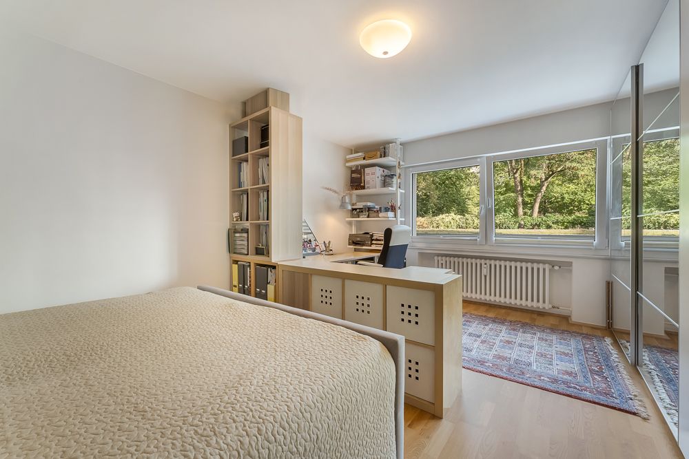 Helles Schlafzimmer mit Schreibtisch, Regal, Teppich und großen Fenstern mit Blick ins Grüne.