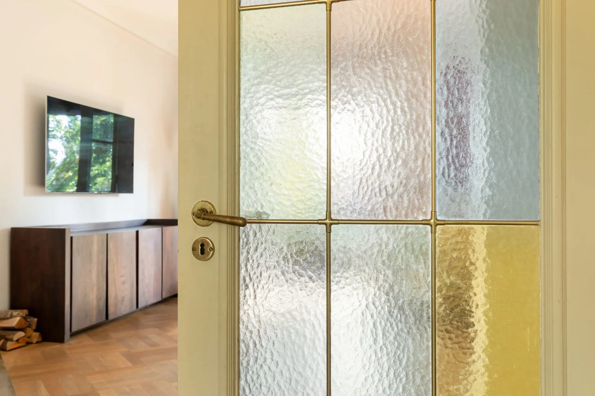 Tür mit strukturiertem Glas, dahinter ein Wohnzimmer mit TV und Holzkommode.