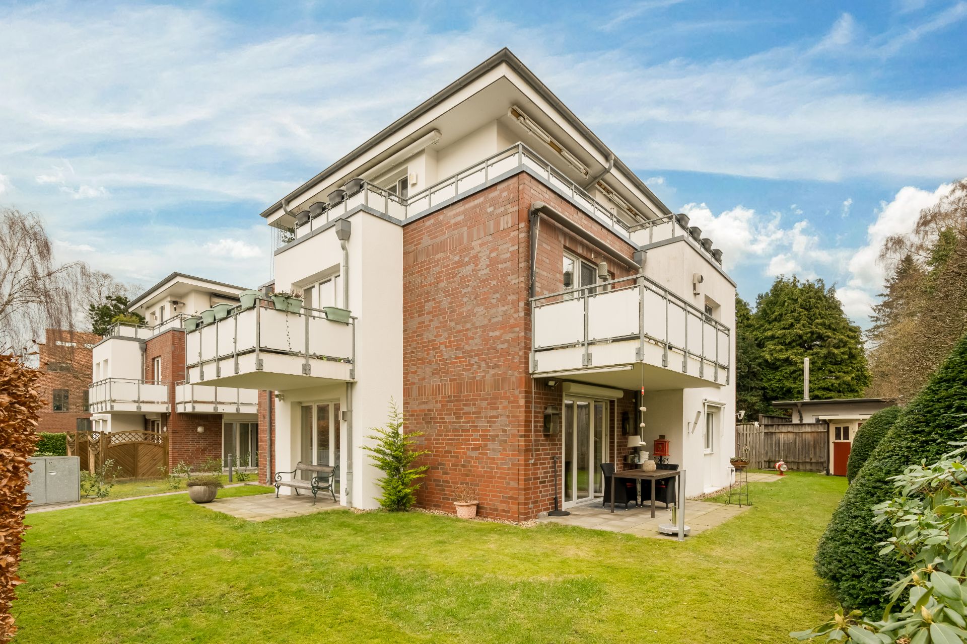 Modernes Wohngebäude mit Balkonen, Terrasse und Garten, umgeben von Rasen und Bäumen.