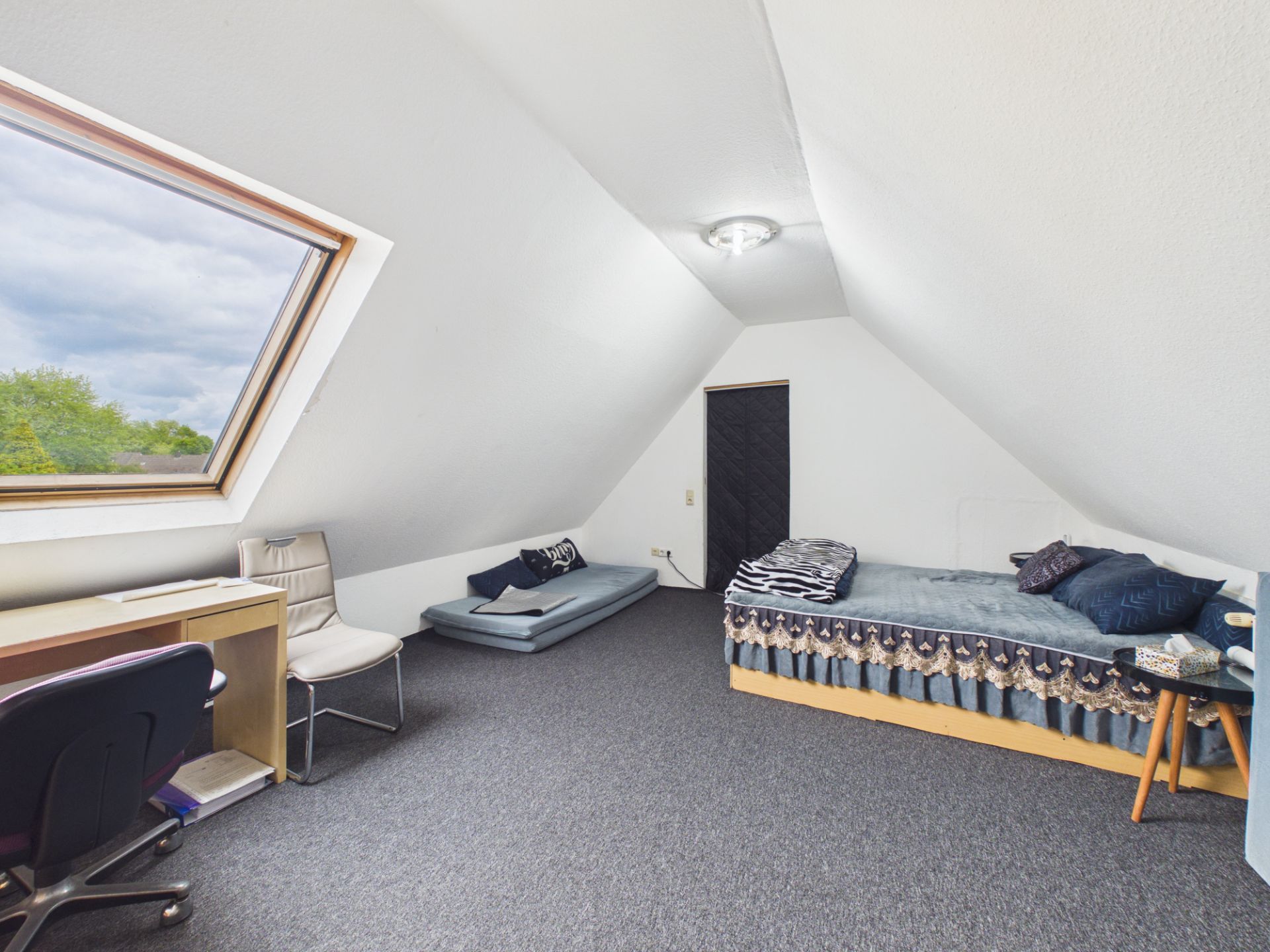 Dachzimmer mit Einzelbett, Schreibtisch, Stuhl und großem Dachfenster.