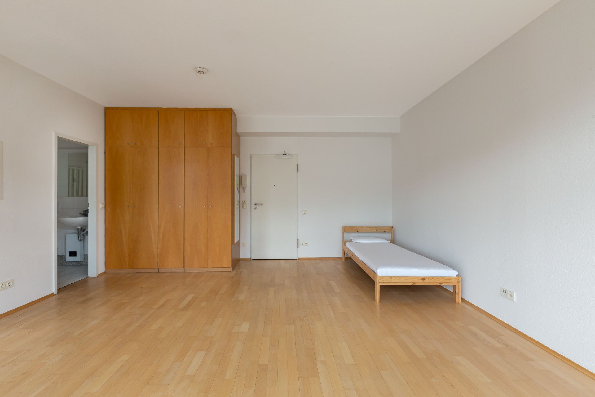 Helles Zimmer mit Holzboden, Einzelbett, Kleiderschrank und Zugang zu einem Badezimmer.