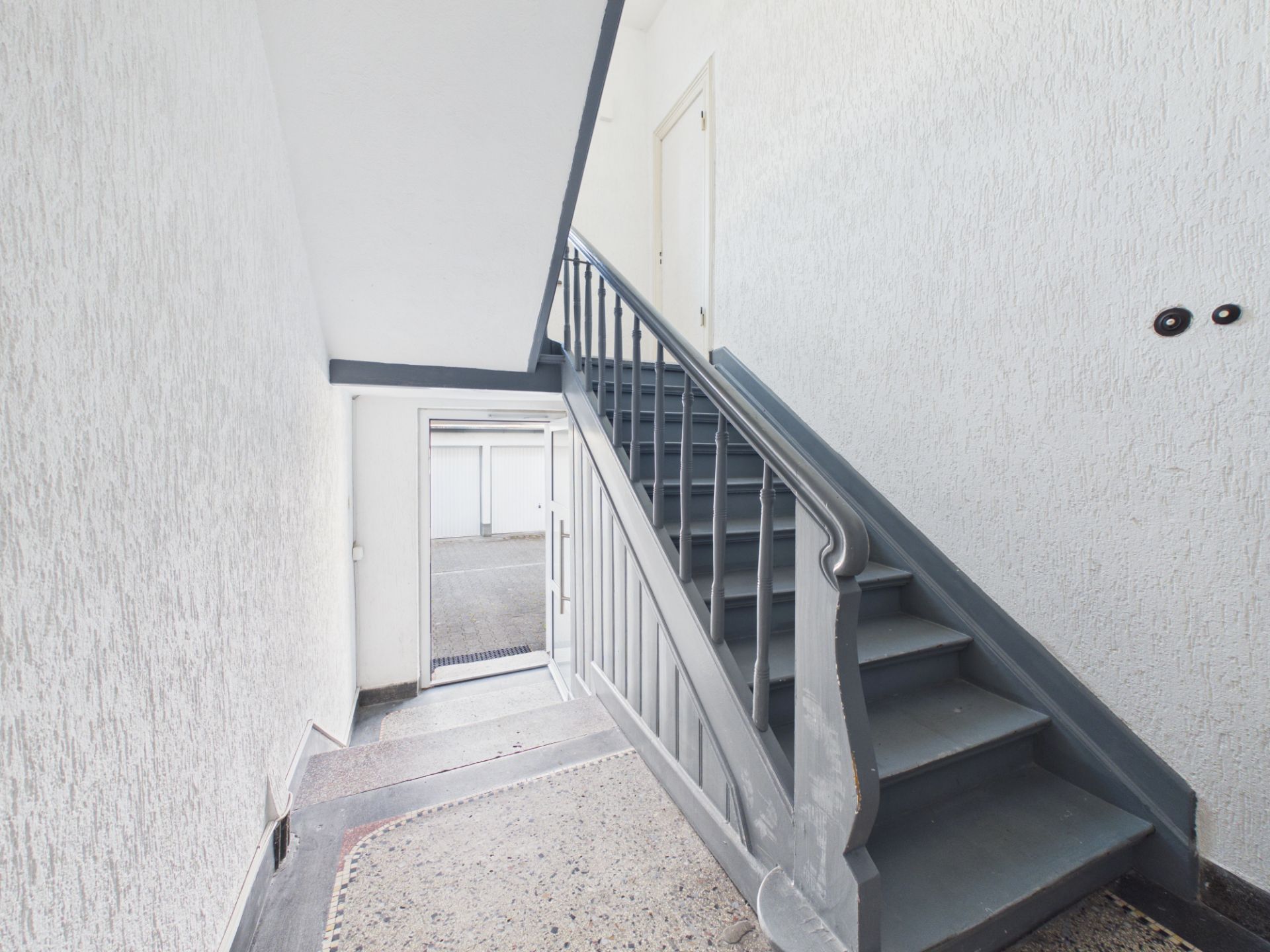 Treppenhaus mit grauer Treppe und weißer Wand, Blick auf Ausgangstür.