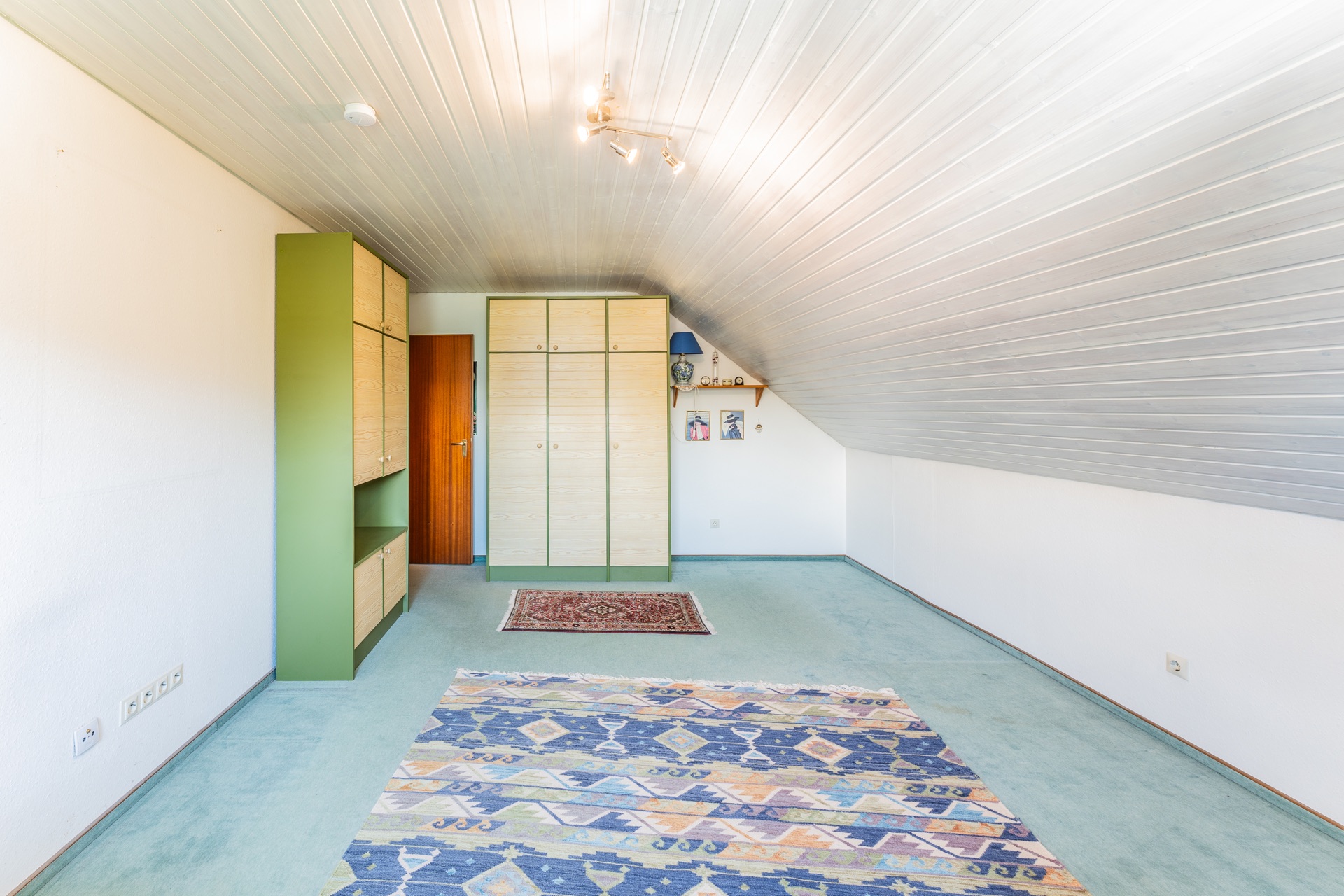 Helles Dachzimmer mit Teppichen, Einbauschränken und Regalen unter der Schräge.