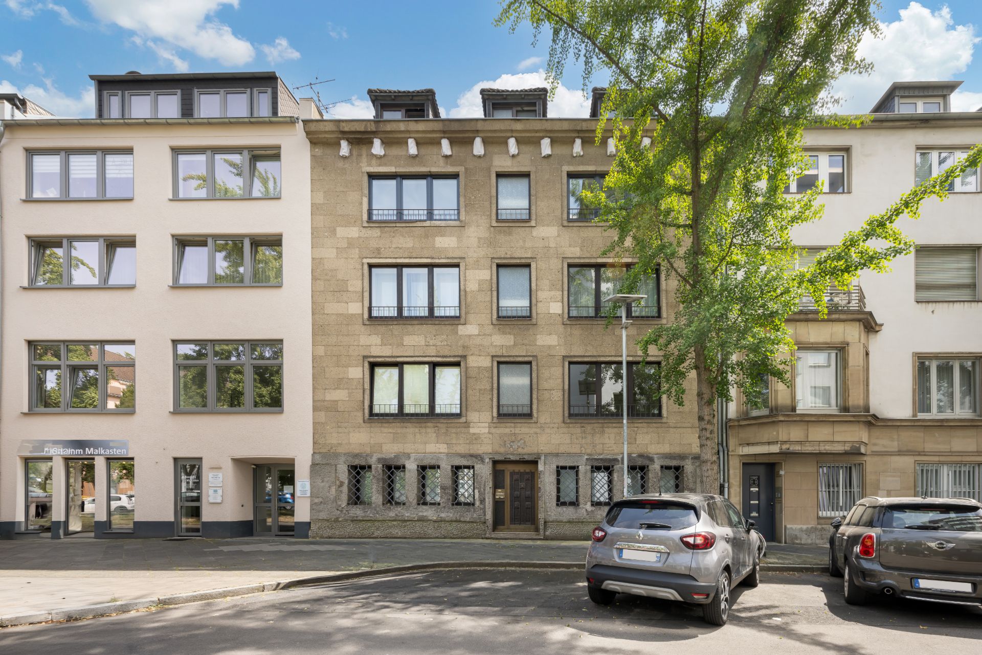 Mehrstöckiges Wohnhaus mit großen Fenstern und einem Baum davor, zwei Autos parken auf der Straße.