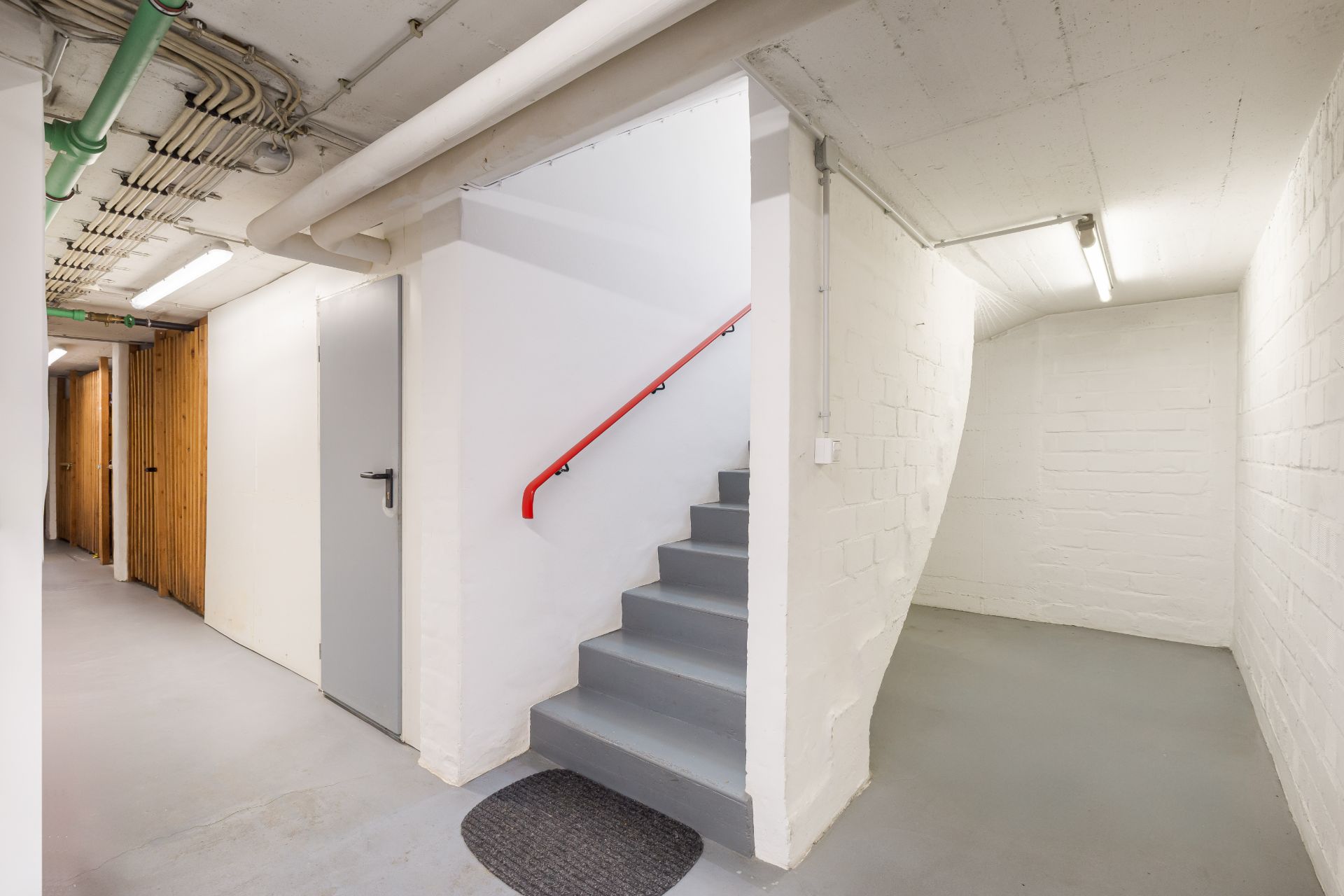 Heller Kellerflur mit Treppe, Türen und sichtbaren Rohren an der Decke.