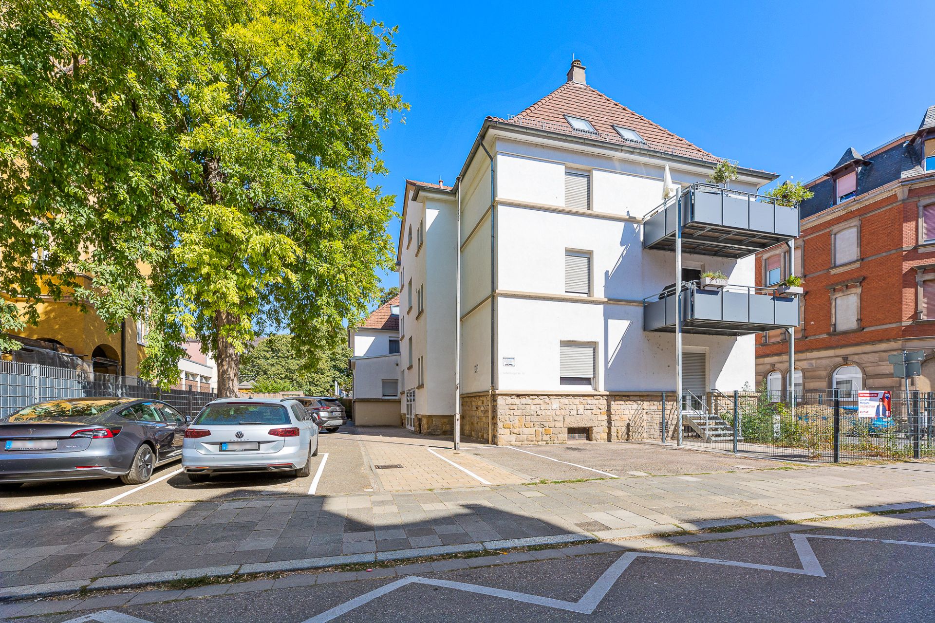 Modernes Wohnhaus mit zwei Balkonen und angrenzendem Parkplatz an einer ruhigen Straße.