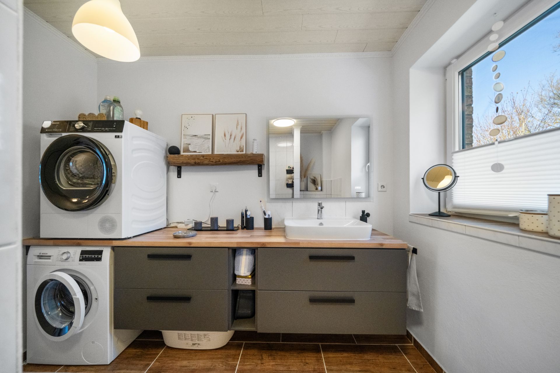 Modernes Badezimmer mit Waschmaschine, Trockner, Waschbecken und großem Spiegel am Fenster.