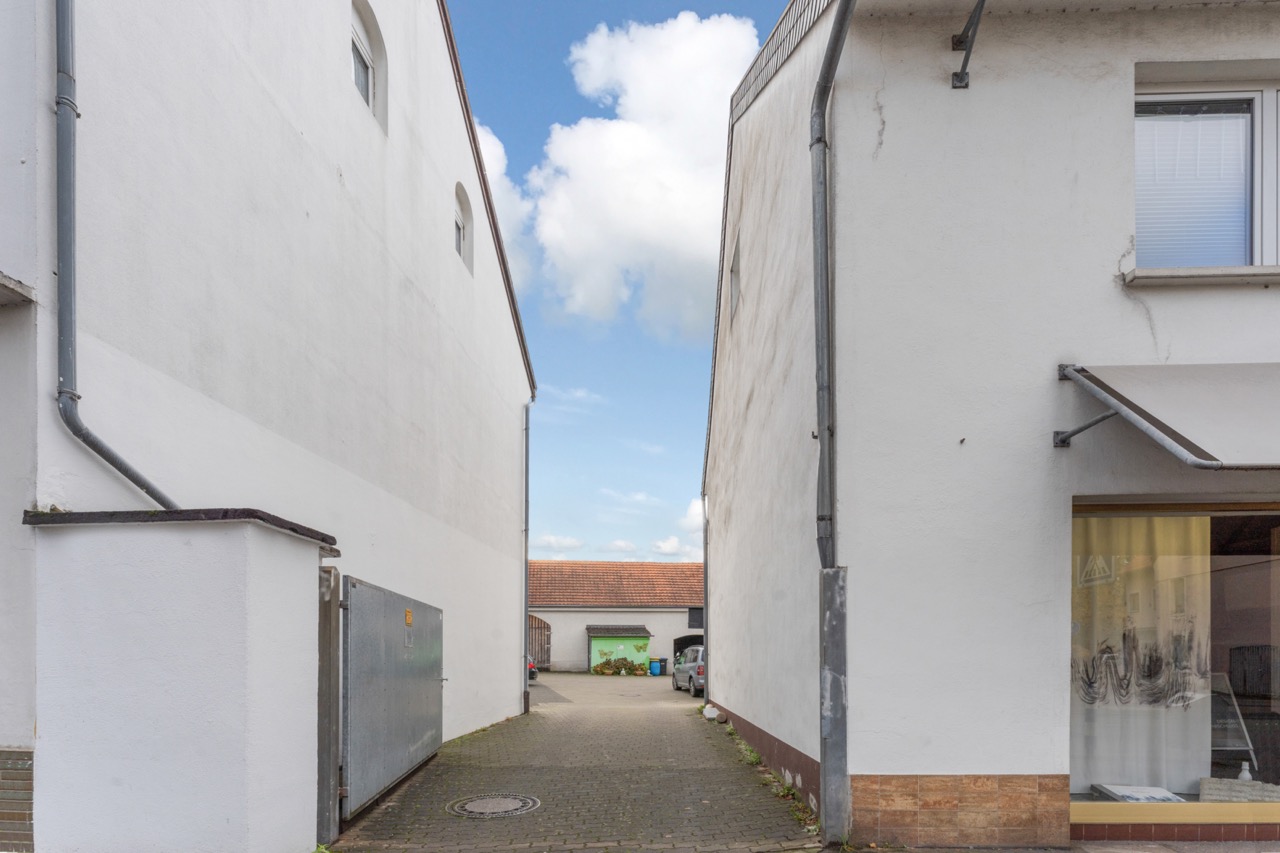 Schmaler Durchgang zwischen zwei weißen Wohnhäusern, Pflasterweg führt zu einem Hof mit Garage.