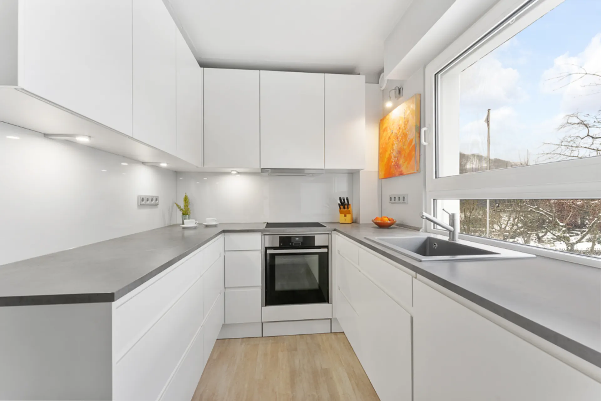 Moderne weiße Küche mit grauer Arbeitsplatte, großem Fenster und Einbauherd.