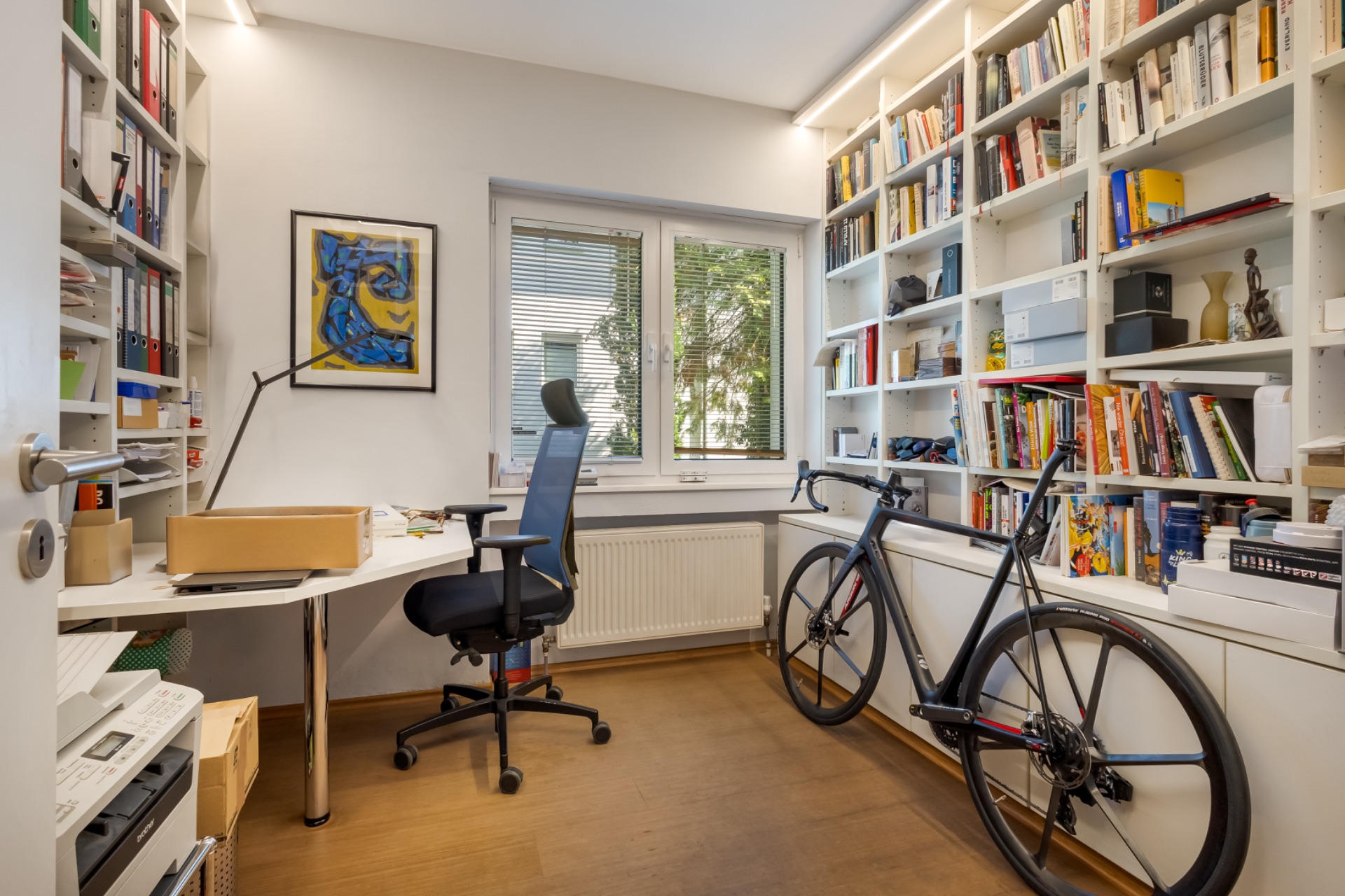 Helles Arbeitszimmer mit Schreibtisch, Drehstuhl, Bücherregal und Fahrrad am Fenster.