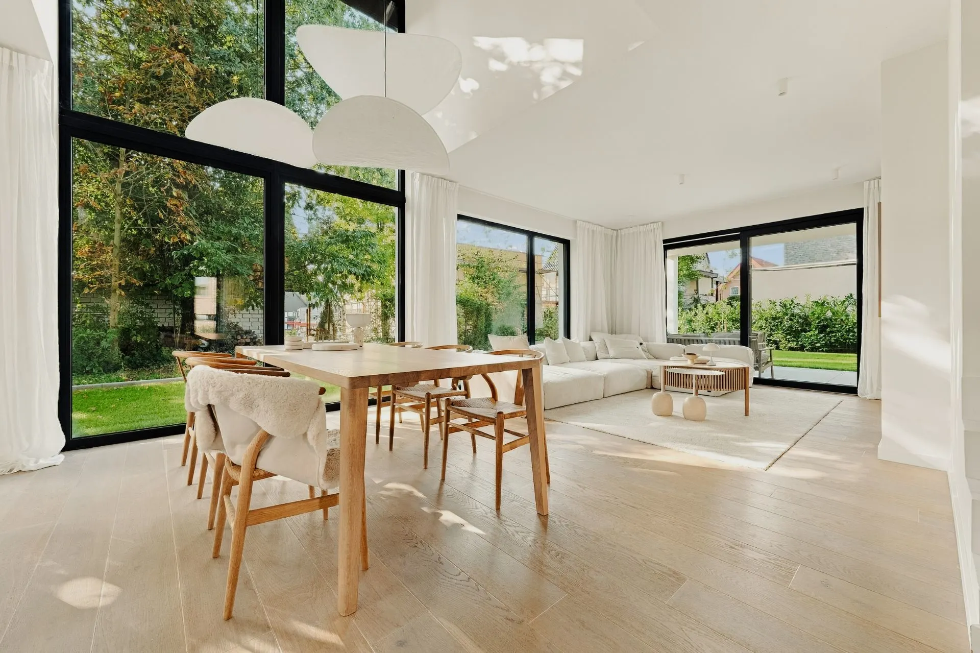 Helles Wohnzimmer mit großem Esstisch, Holzstühlen und Sofa, große Fenster mit Blick auf Garten.