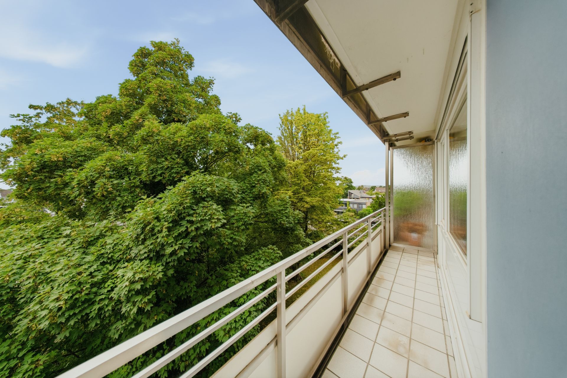 Langer Balkon mit weißen Fliesen und Geländer, Blick auf grüne Bäume.