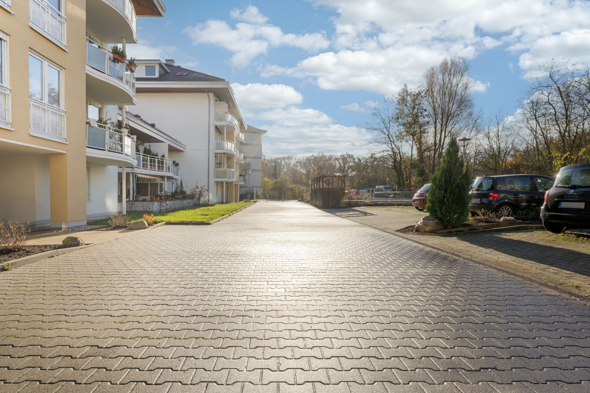 Gepflasterter Weg zwischen modernen Wohngebäuden mit Balkonen und parkenden Autos rechts.