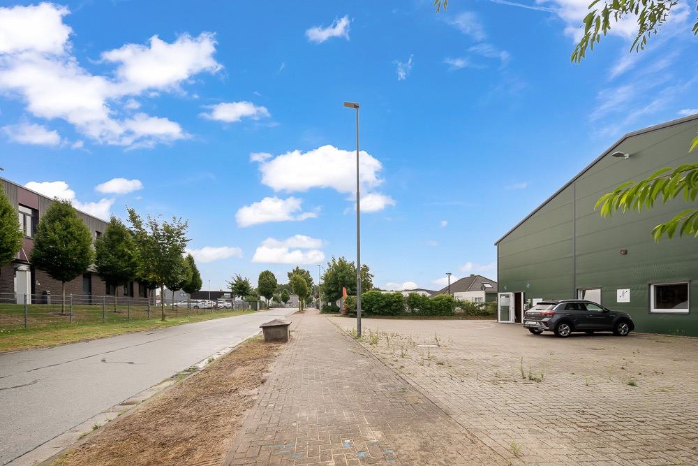 Gehweg mit Bäumen links, rechts ein Parkplatz und ein großes grünes Gebäude unter blauem Himmel.