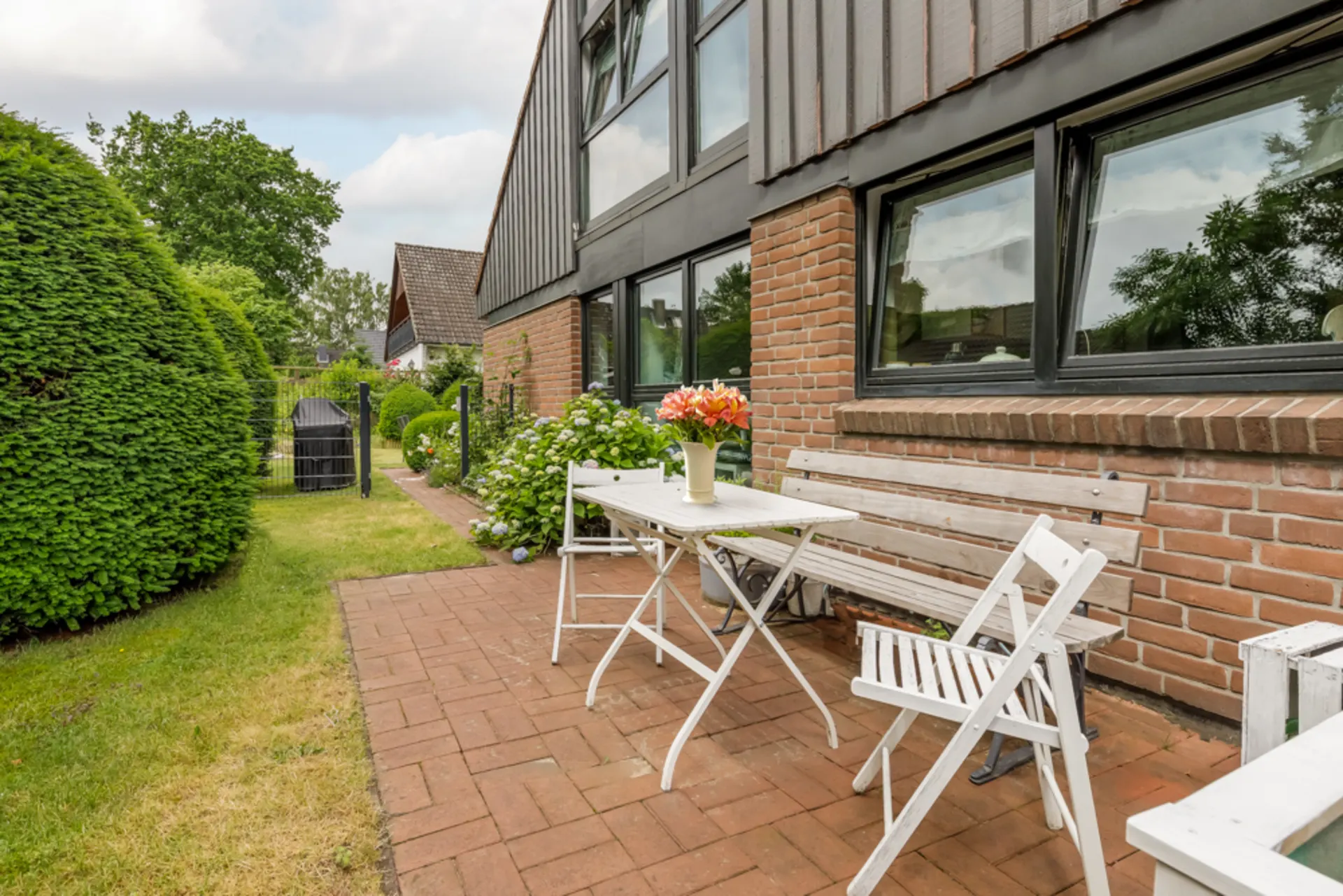 Terrasse mit weißer Sitzbank, Stühlen und Tisch, darauf ein Blumenstrauß, neben einem Backsteinhaus.