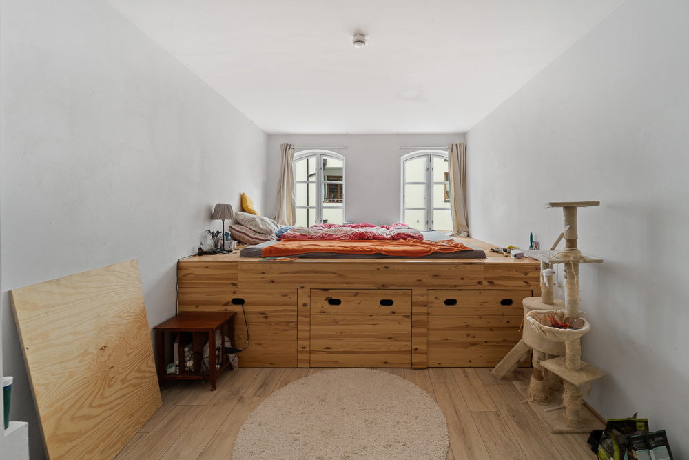 Schlafzimmer mit erhöhtem Holzbett, Katzenturm und zwei Fenstern mit hellen Vorhängen.