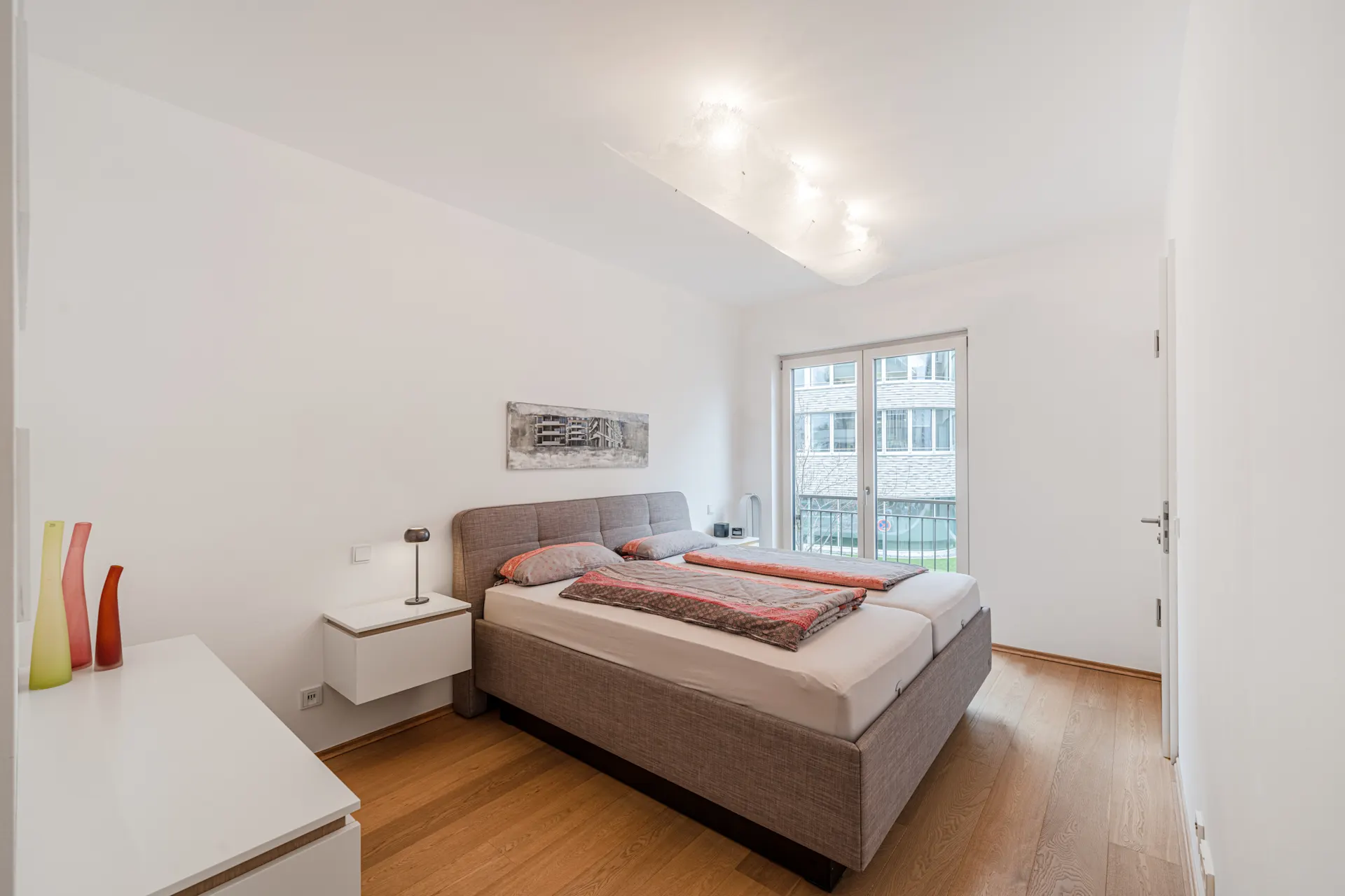 Helles Schlafzimmer mit Doppelbett, Nachttischen, Kommode und großem Fenster.