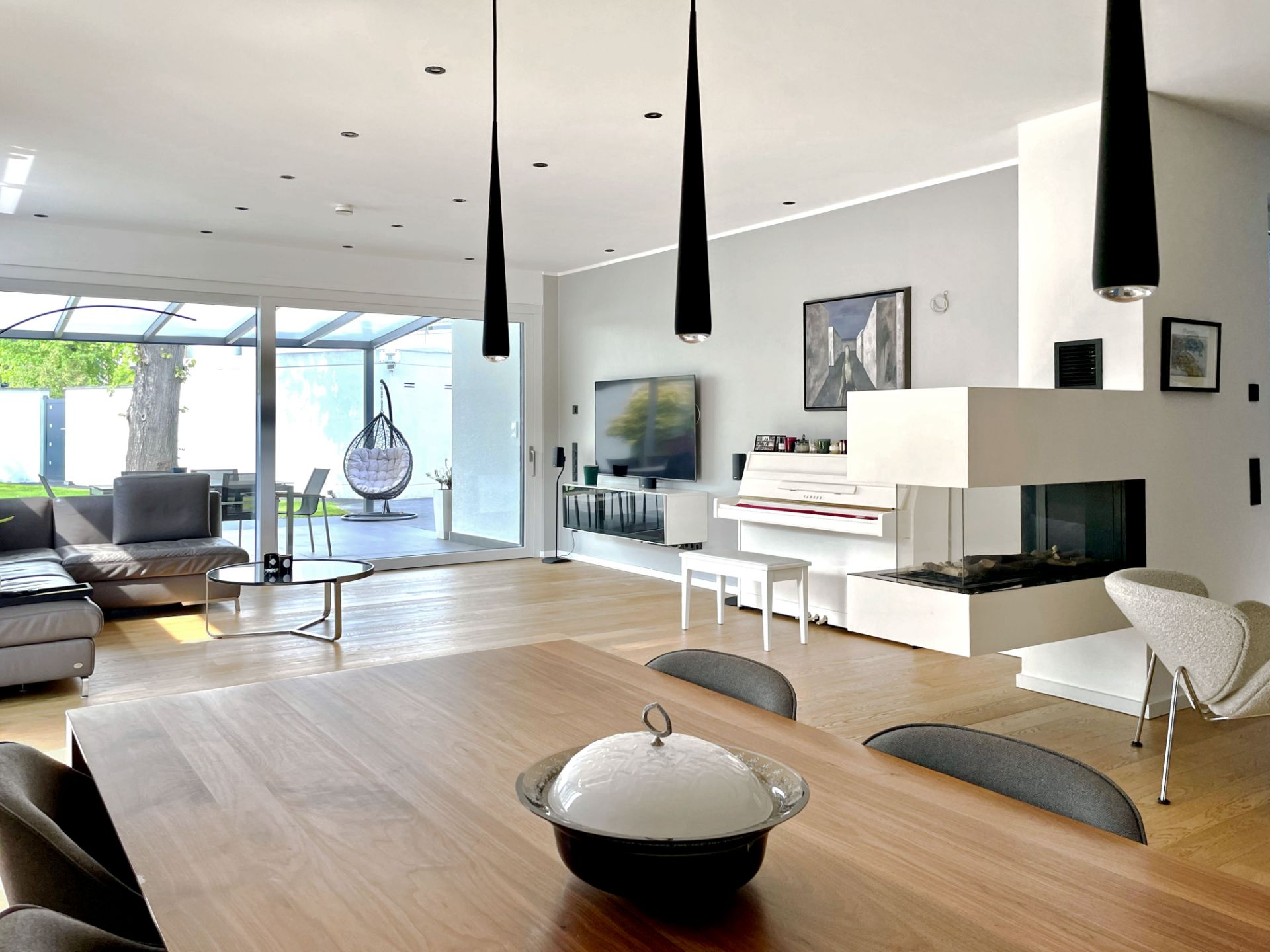 Helles Wohnzimmer mit Kamin, Sofa, Essbereich und großer Glasfront zum Garten.