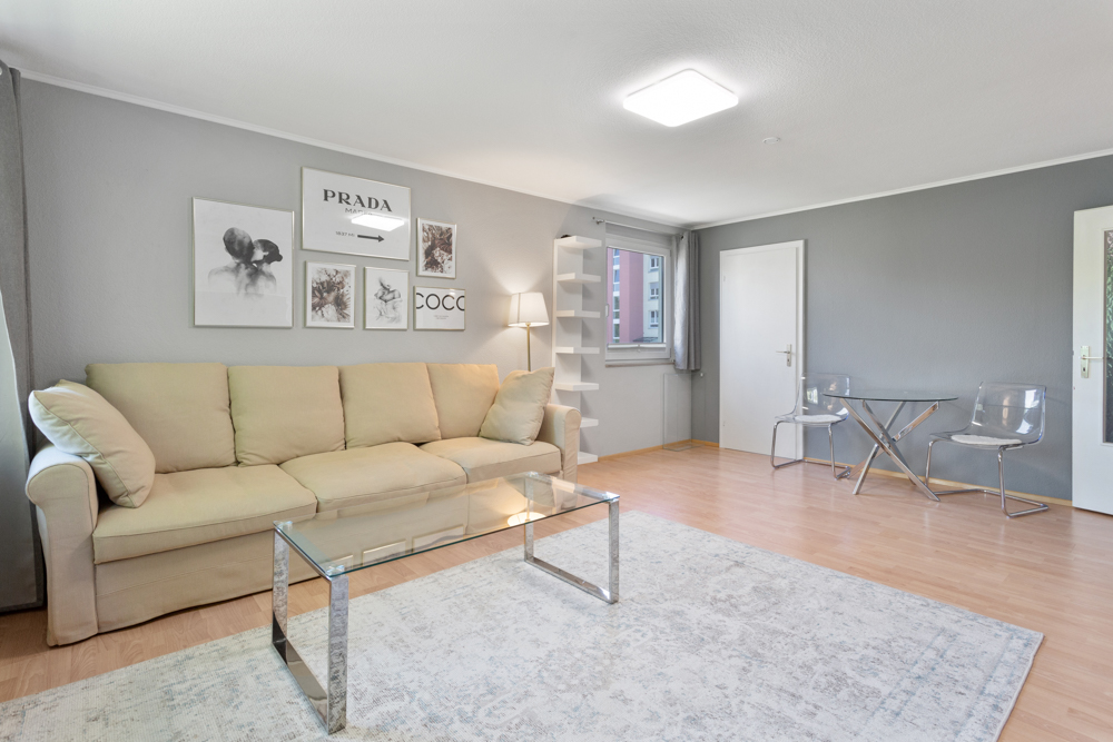 Helles Wohnzimmer mit beiger Couch, Glastisch, Teppich und kleinem Esstisch mit zwei Stühlen.