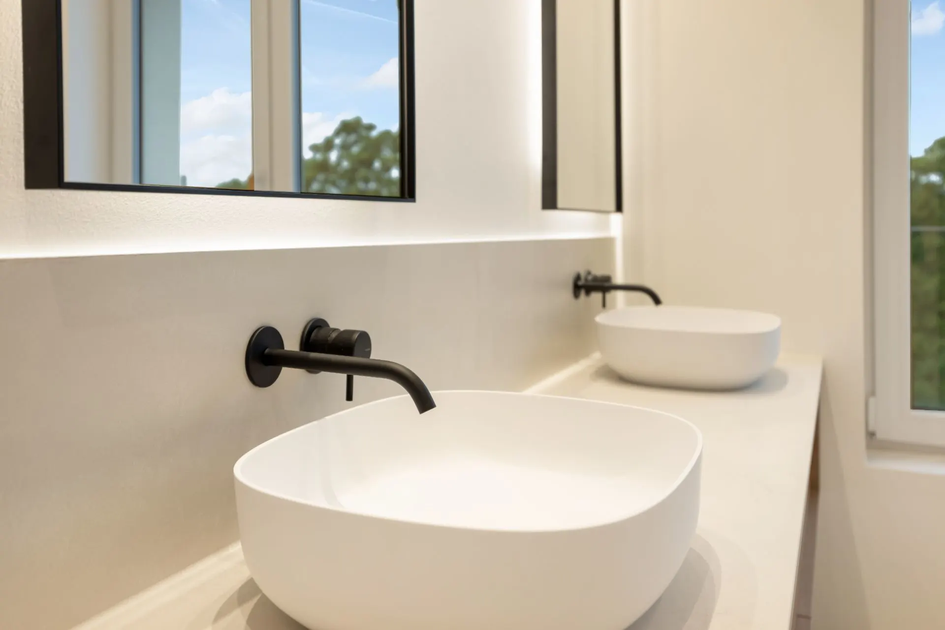 Zwei moderne, weiße Waschbecken mit schwarzen Wasserhähnen vor Spiegeln in einem hellen Bad.