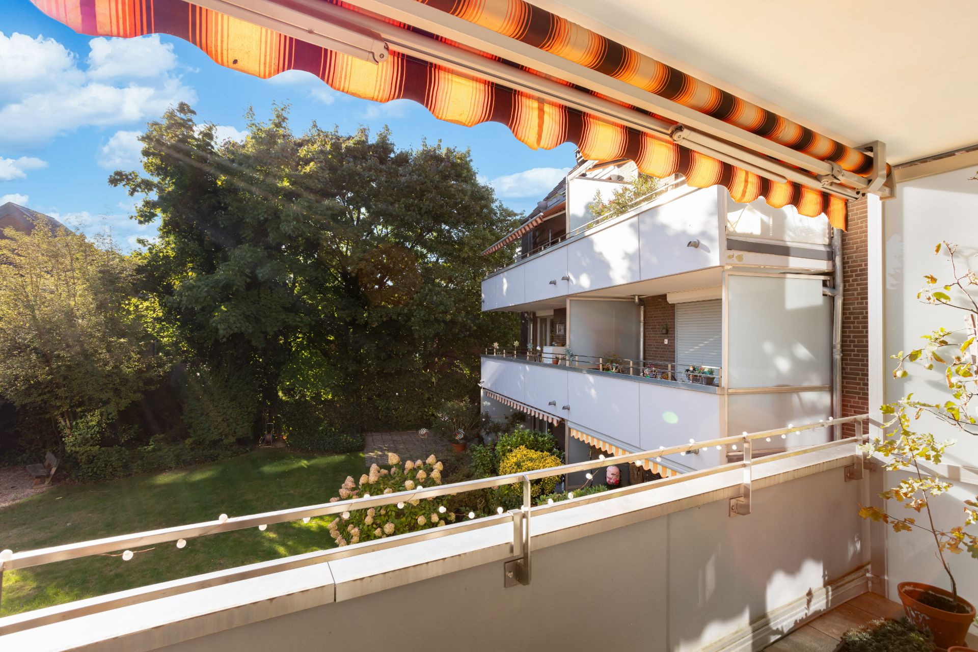 Blick von einem Balkon mit Markise auf einen grünen Garten und Nachbarbalkone.