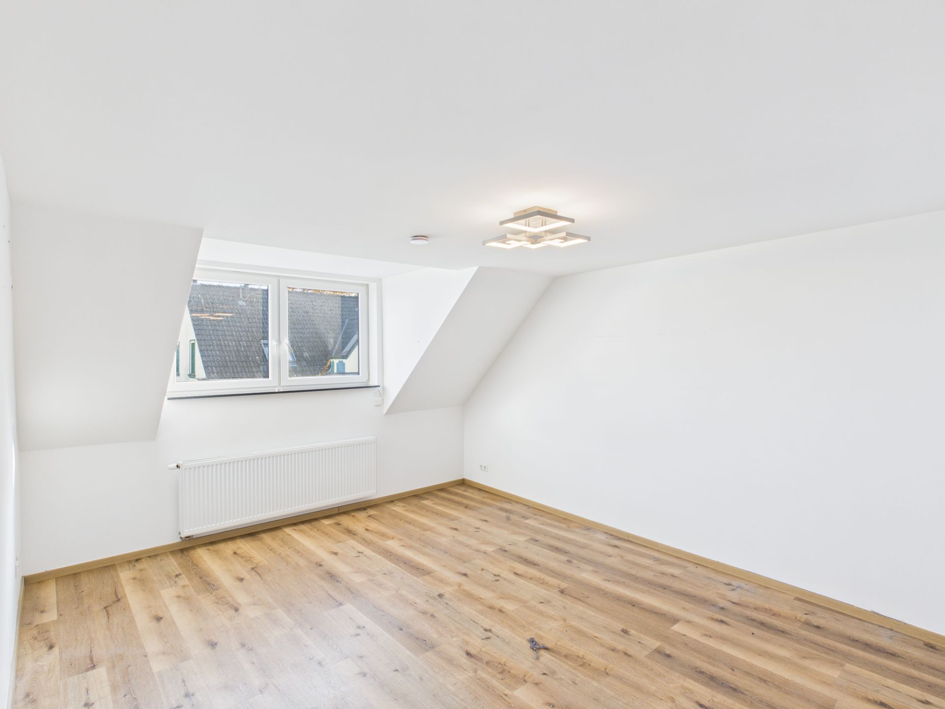 Heller Raum mit Dachschräge, Holzfußboden, Fenster und Heizkörper unter dem Fenster.