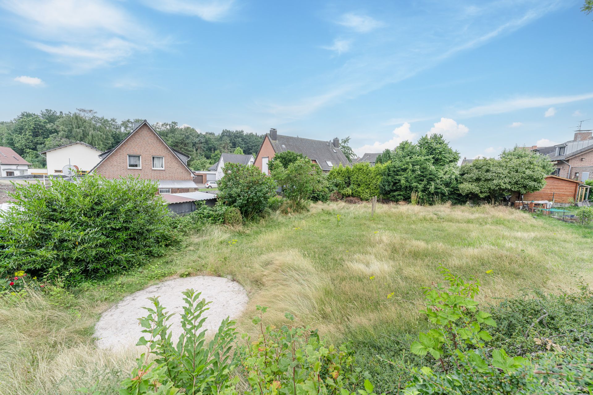 Grünes Grundstück mit hohem Gras, Büschen und mehreren Einfamilienhäusern im Hintergrund.