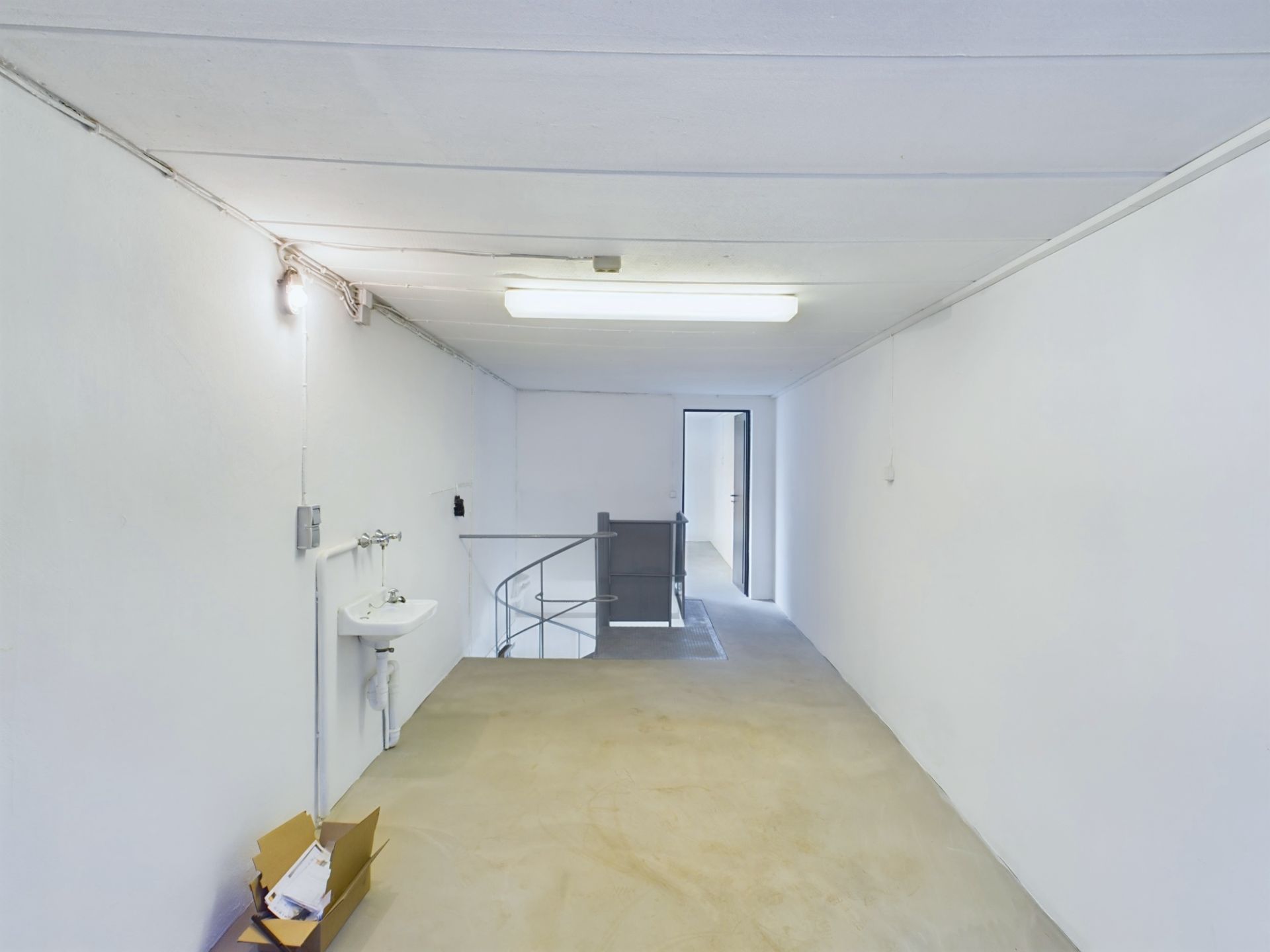 Weißer Kellerraum mit Waschbecken, Karton und Zugang zu einer Wendeltreppe.