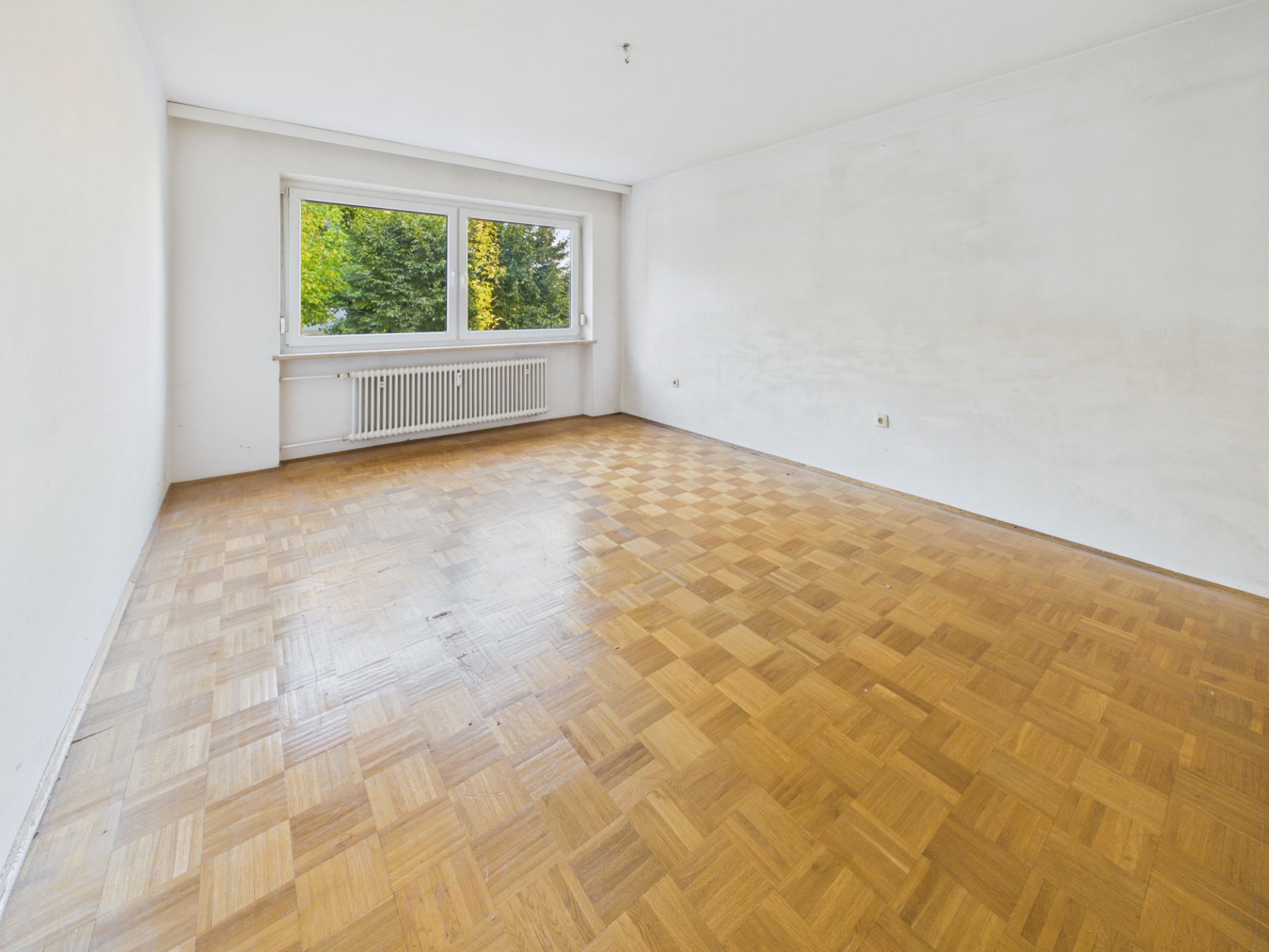 Leerer Raum mit Parkettboden, weißer Wand und großem Fenster mit Blick ins Grüne.